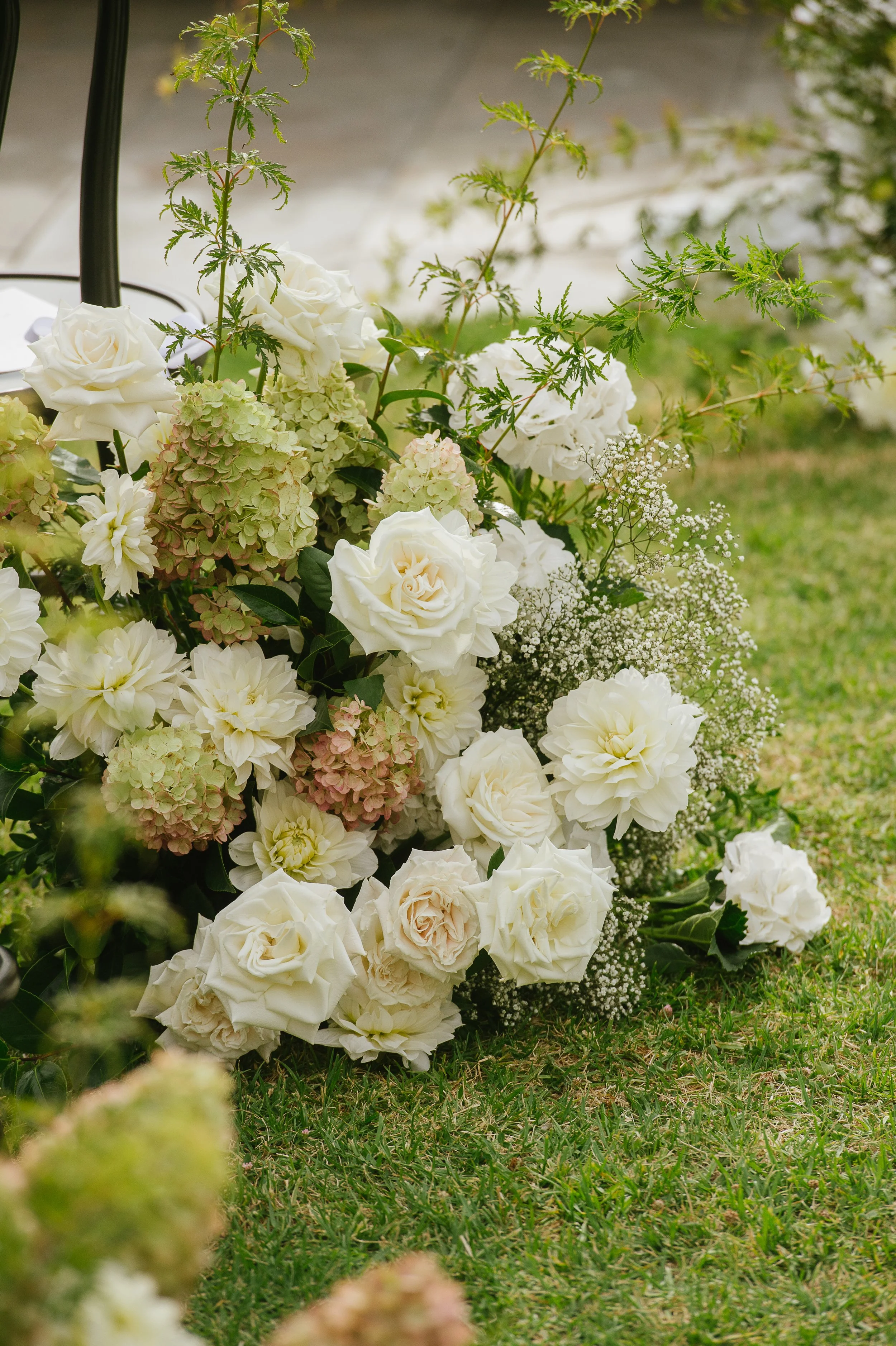 A floral arrangement consisting of white roses, dahlias, hydrangeas, baby's breath, and greenery resting on grass outside.