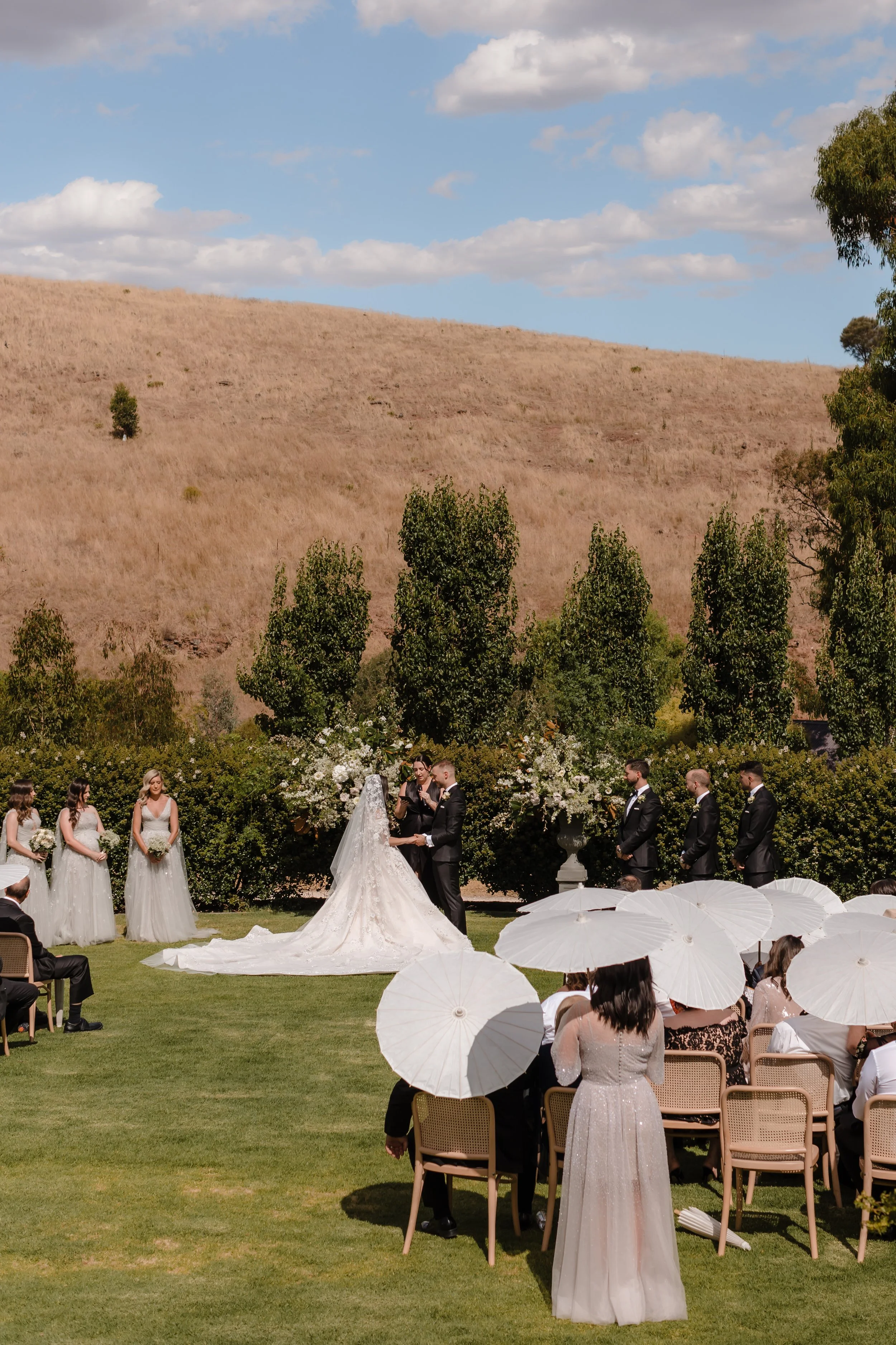 A wedding ceremony outdoors with the bride and groom at the altar, surrounded by bridesmaids and groomsmen, with guests seated under white umbrellas on a grassy lawn and a hillside with trees in the background.