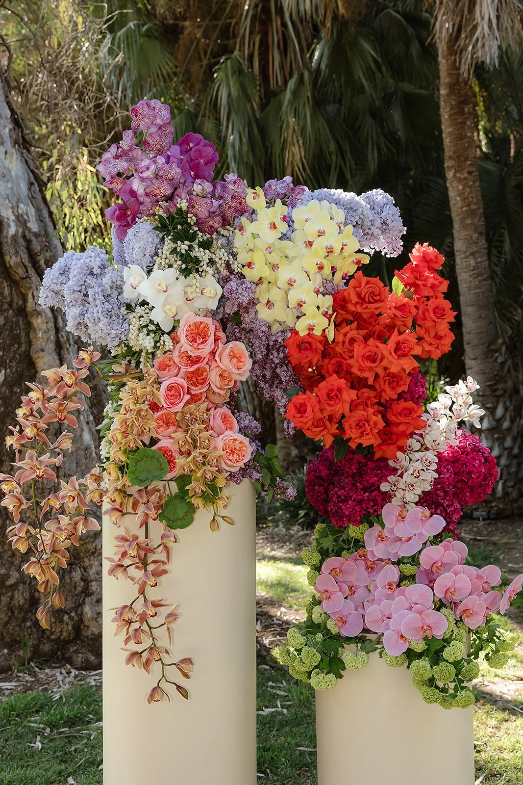 Two tall beige vases filled with colorful flowers including orchids, roses, hydrangeas, and other blooms, set outdoors with trees and palm fronds in the background.