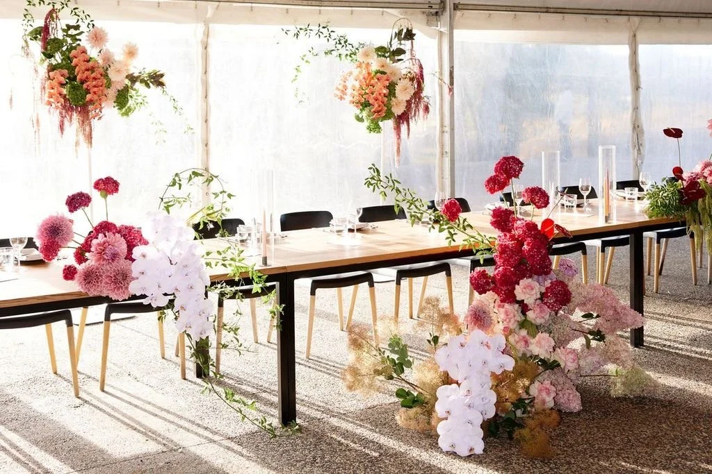 A decorated banquet table with pink, white, and red floral arrangements in a bright, airy tent setup featuring elegant candleholders and black chairs.