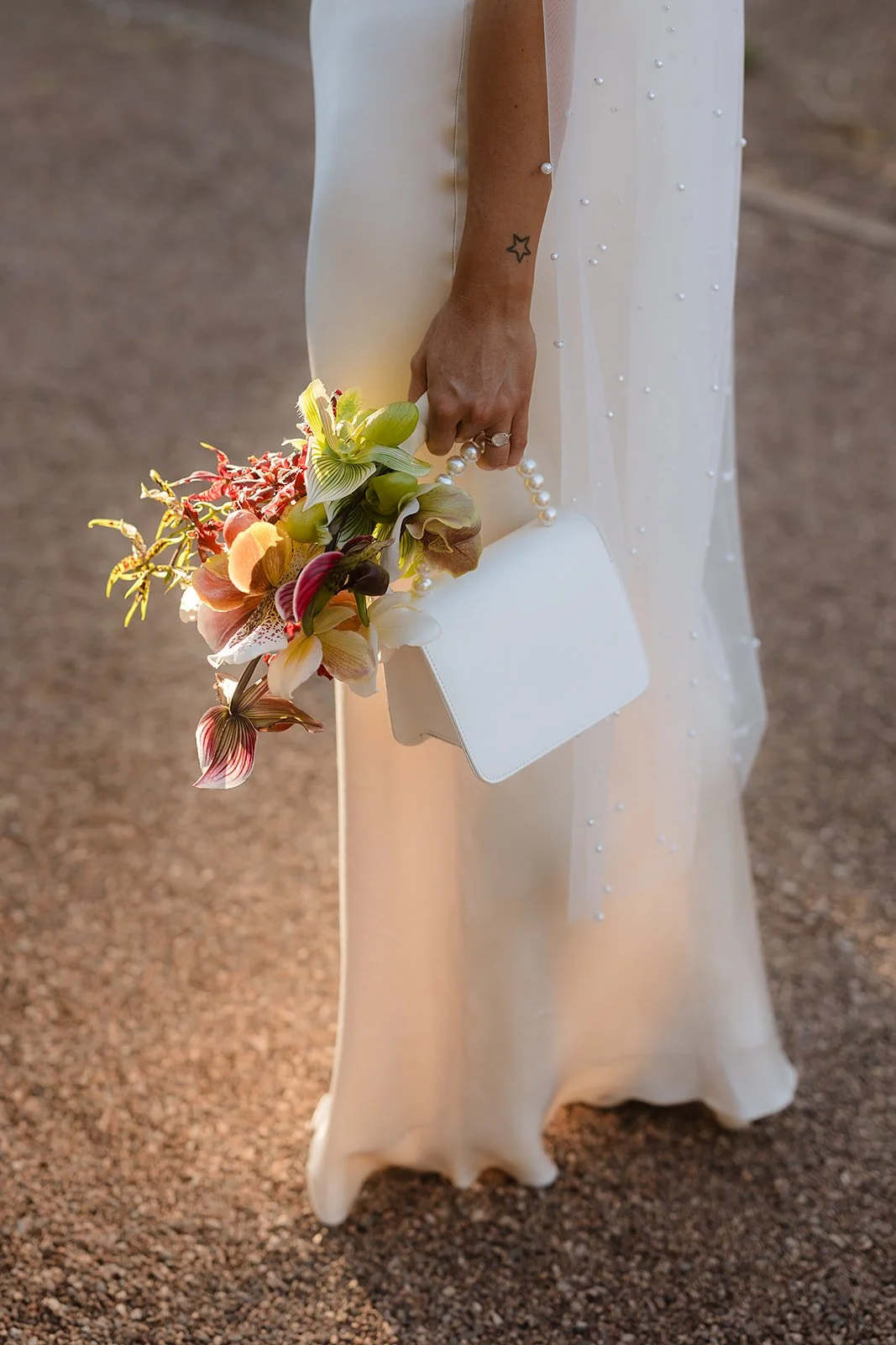 Close-up of a woman's hand holding a white clutch, a pearl necklace, and a bouquet of colorful orchids, with a white dress featuring pearl embellishments.