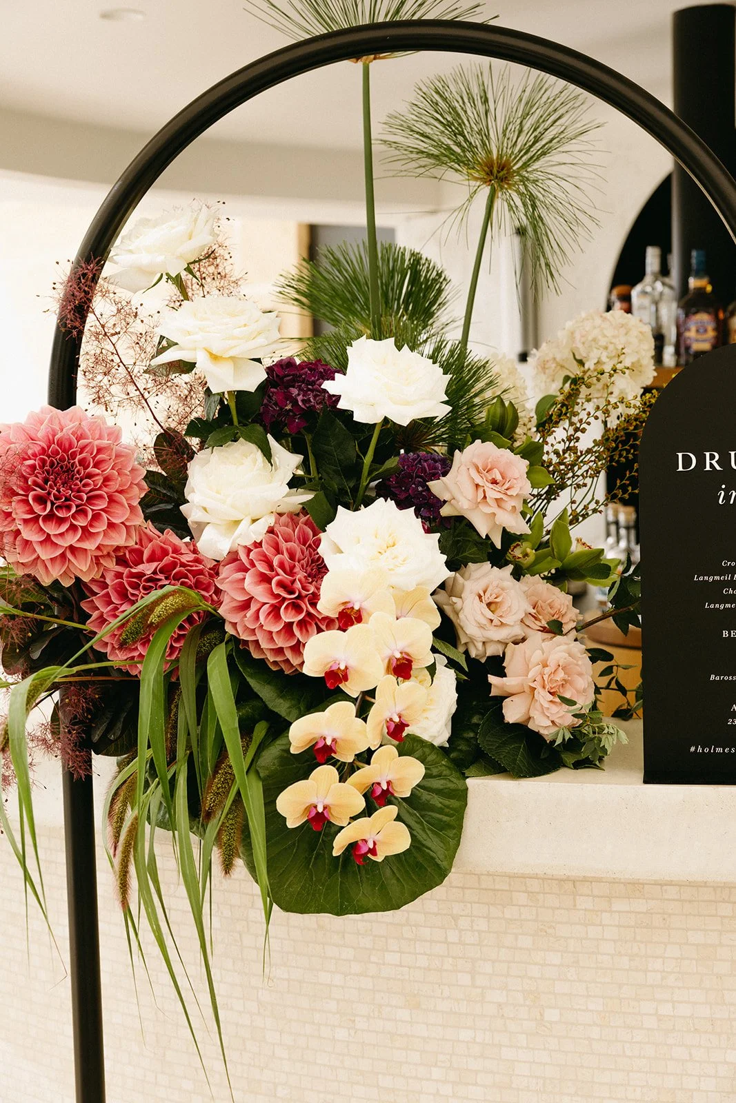 A floral arrangement with white roses, pink dahlias, cream orchids, purple hydrangeas, and green foliage on a beige textured surface, with a black sign partially visible on the right.