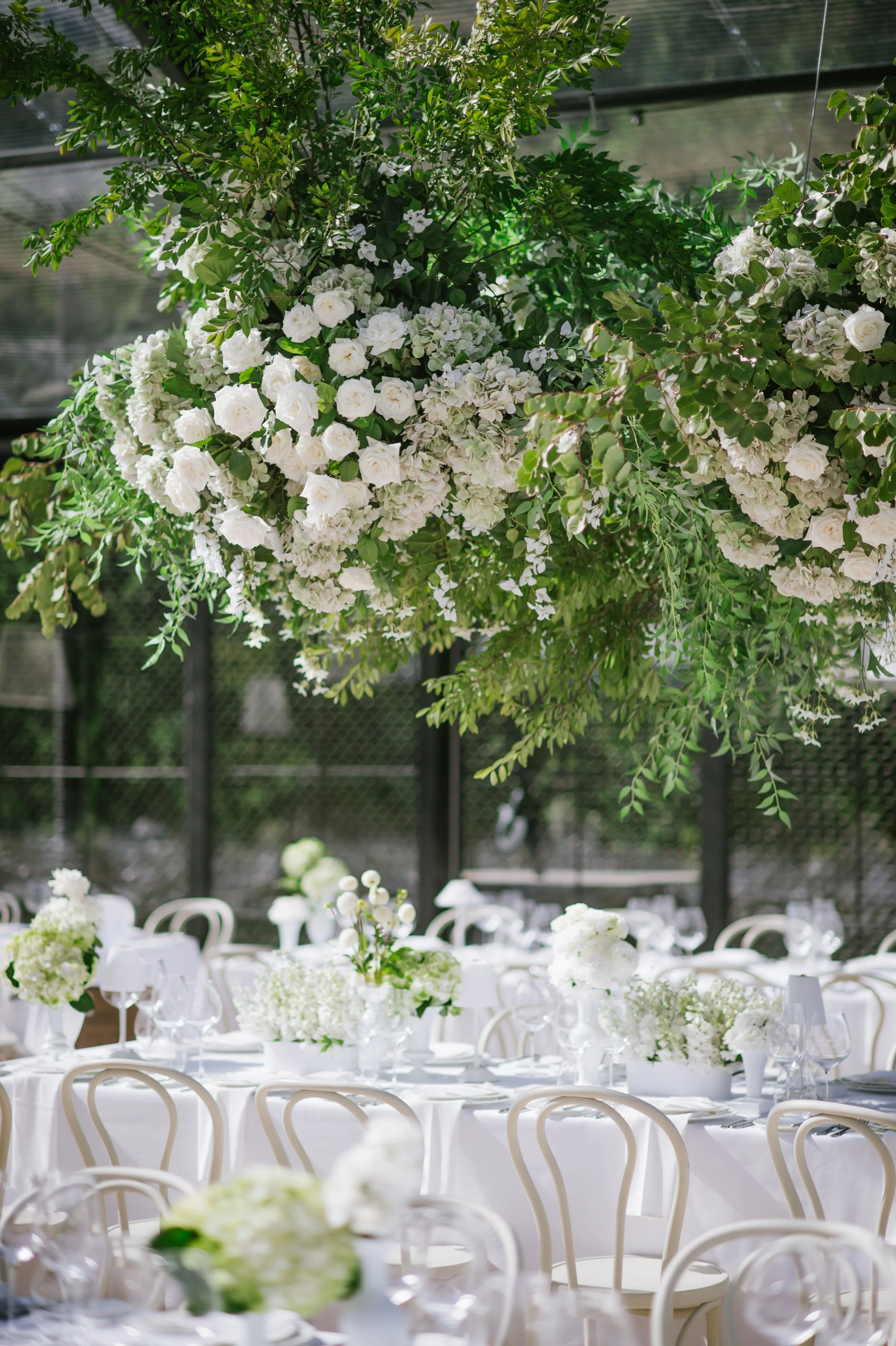 A hanging floral arrangement with white flowers and green leaves over a table set for a wedding or event inside a glass-enclosed space.
