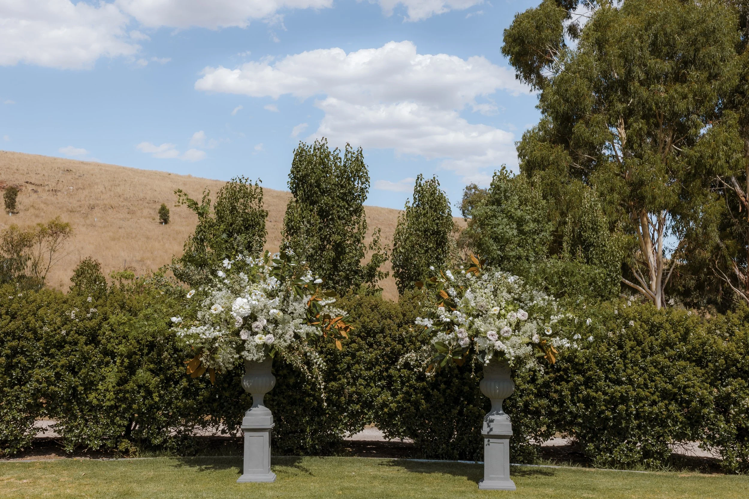 Two large white floral arrangements in white urn vases outdoors, with trees, green bushes, a grassy area, a hill, and a partly cloudy sky in the background.