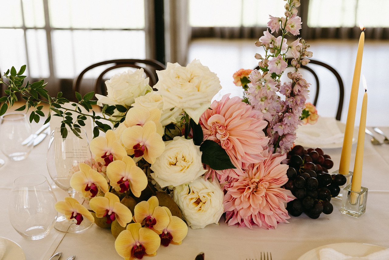A floral centerpiece on a dining table with white, pink, and purple flowers, grapes, and two yellow candles in glass holders.