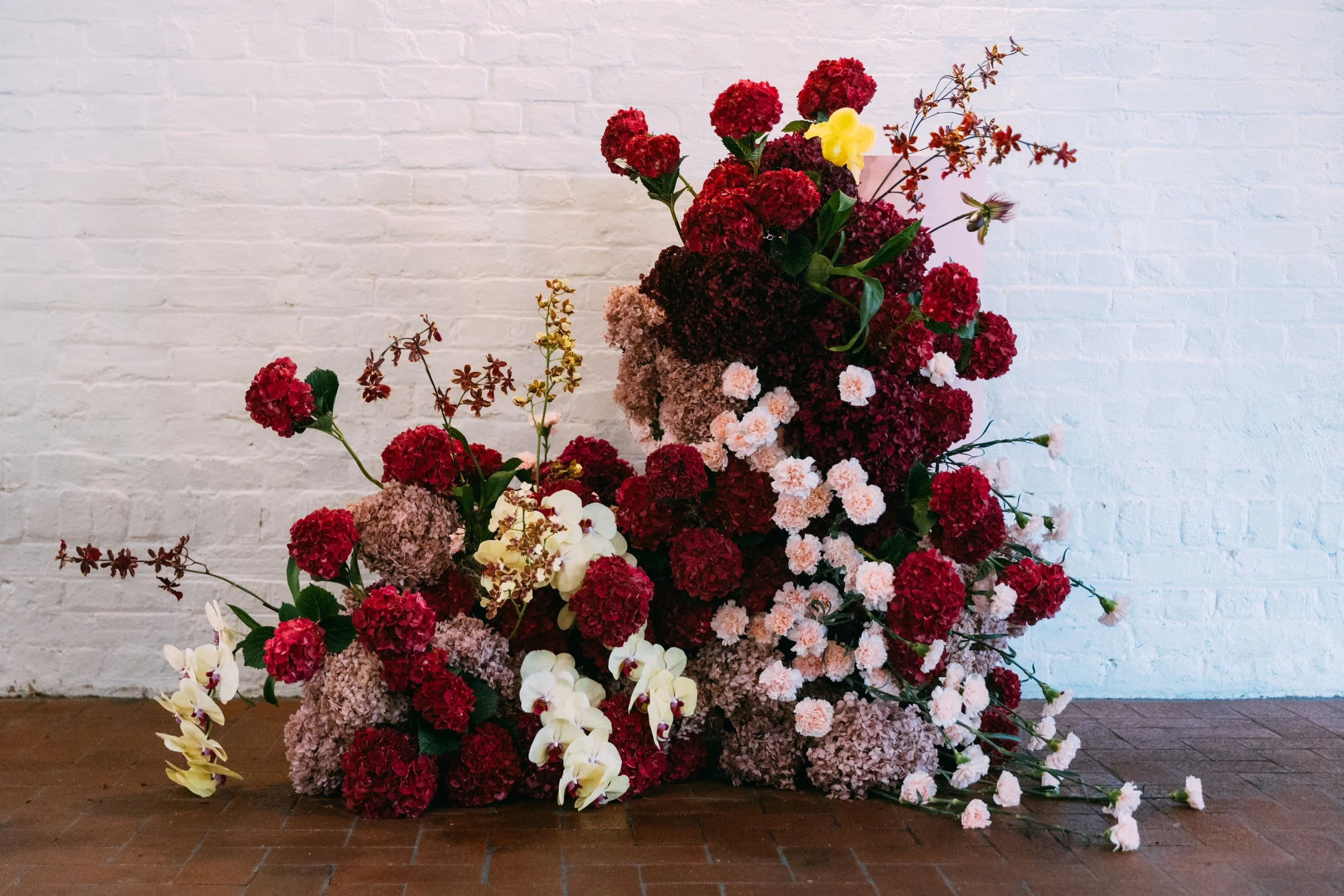 A large arrangement of assorted red, pink, and white flowers, including hydrangeas, orchids, and other blossoms, set against a white brick wall.