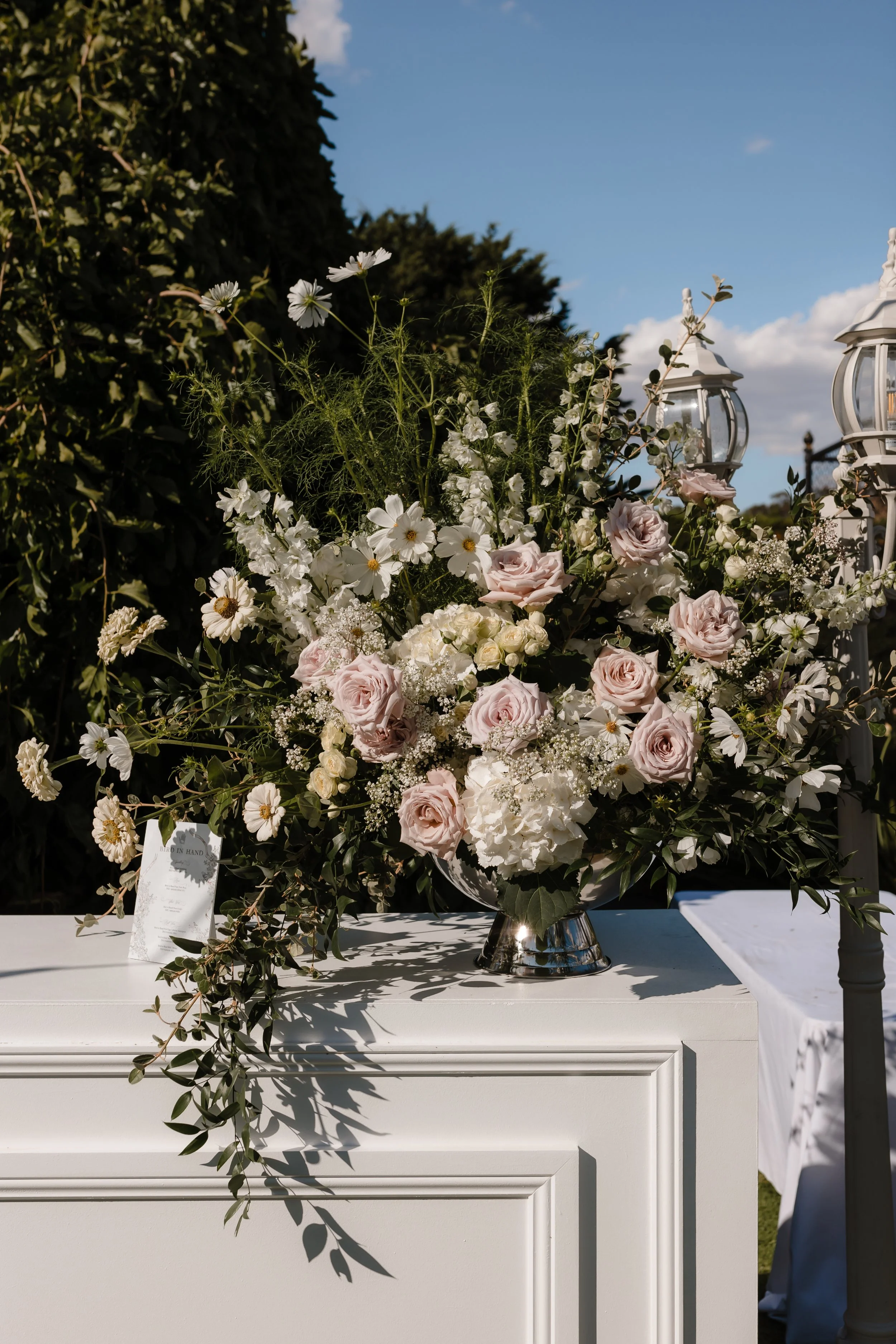 A large floral arrangement with pale pink roses, white hydrangeas, and daisies, placed on a white cabinet outdoors, with two white lanterns nearby and greenery in the background under a partly cloudy sky.