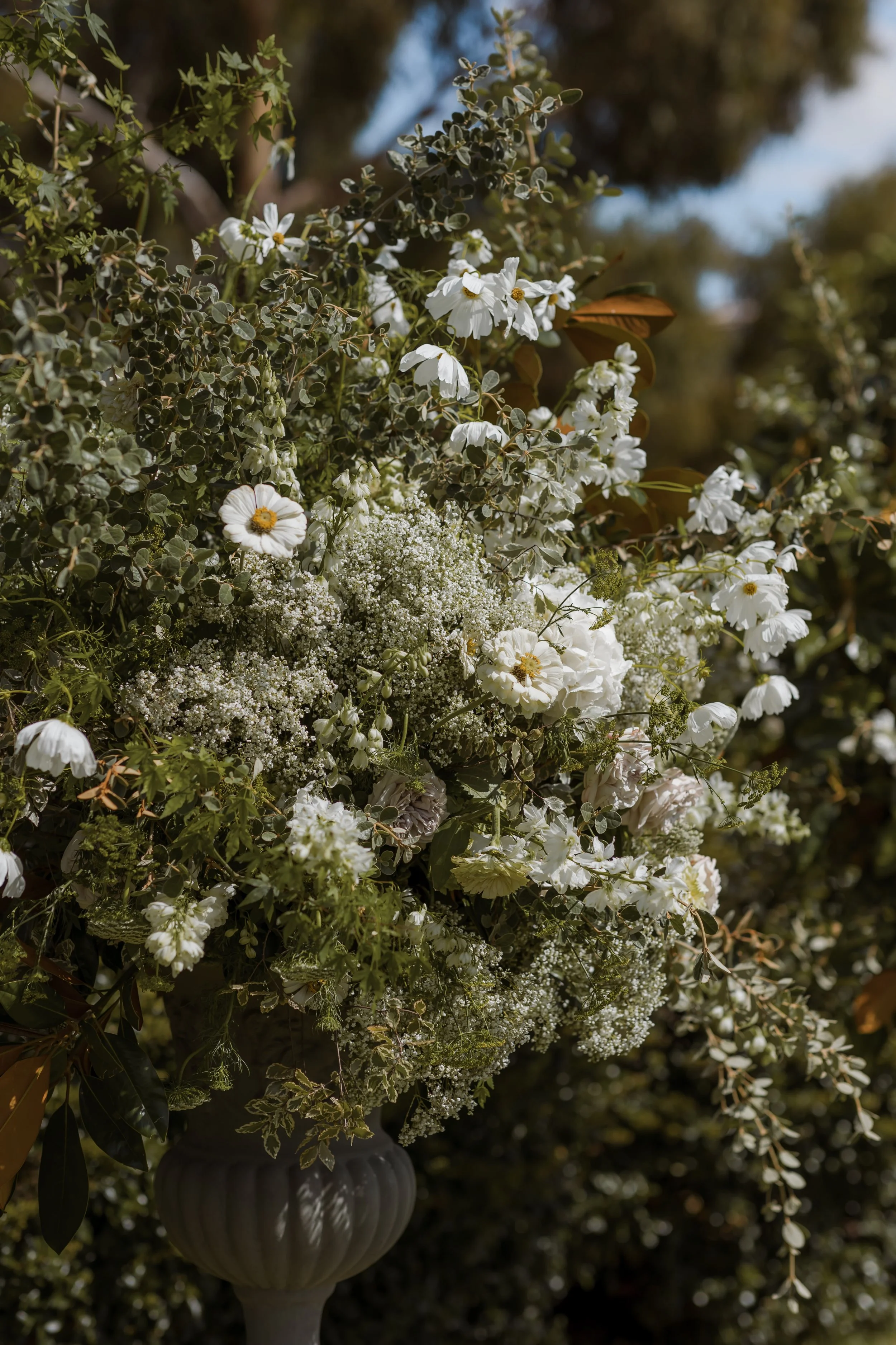 A large flower arrangement with white flowers, including roses and other blooms, in a rounded, dark-colored vase outdoors with trees and sky in the background.
