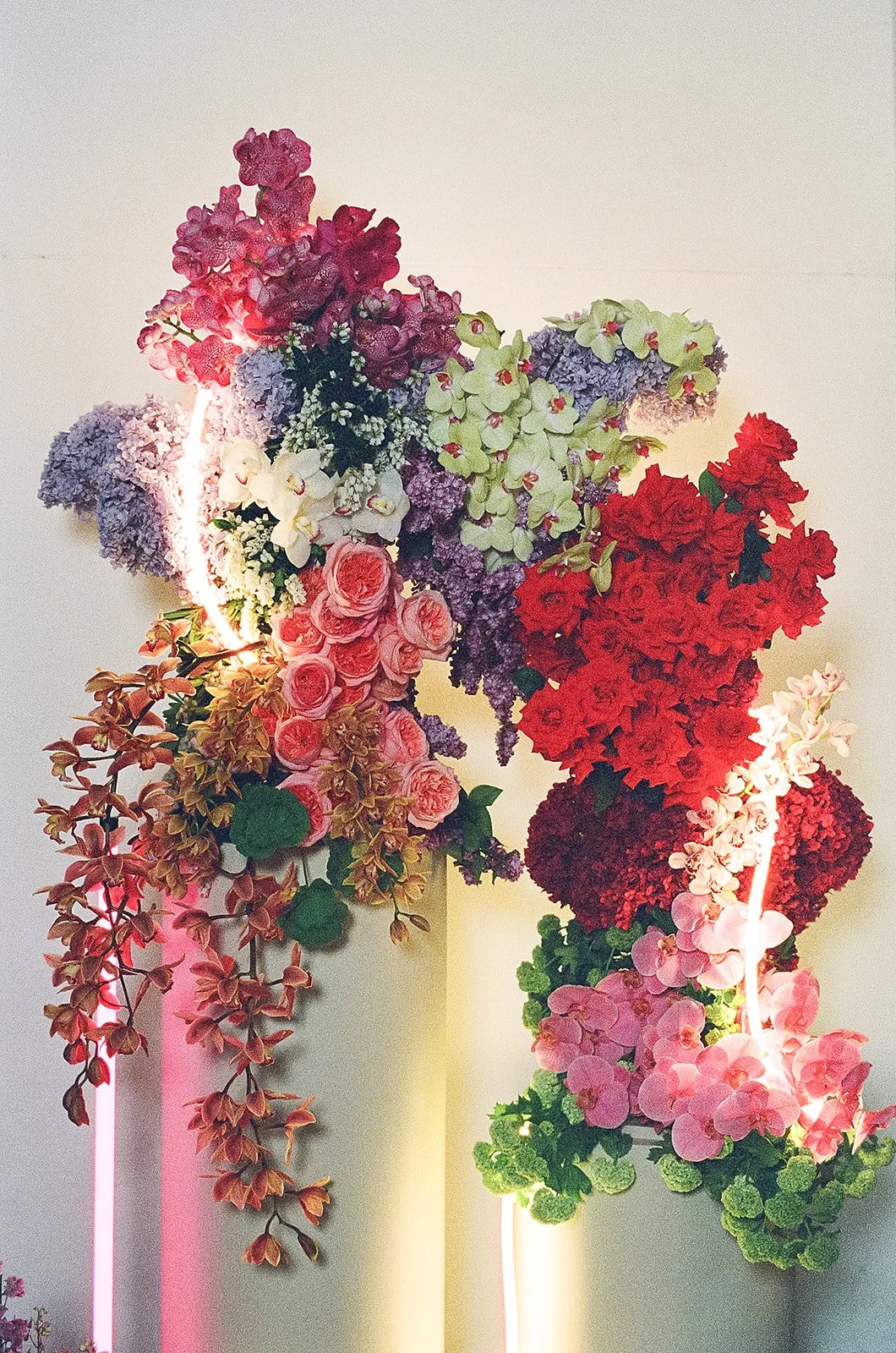 Colorful floral arrangement featuring pink roses, purple and green hydrangeas, red flowers, and cascading greenery, with illuminated pink and white neon lights on a plain light-colored wall.