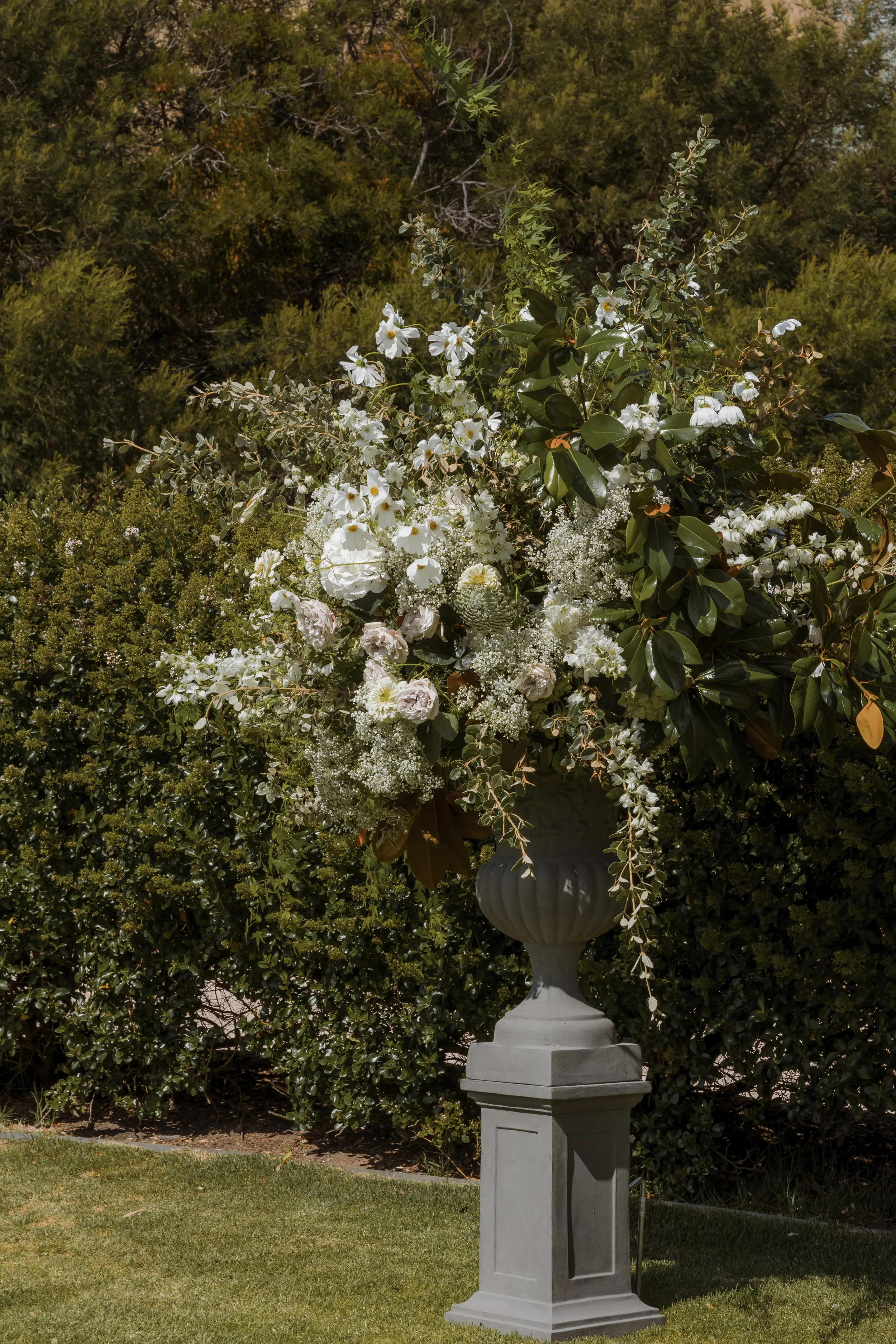 A large floral arrangement in a decorative urn on a pedestal outdoors with green bushes and trees in the background.