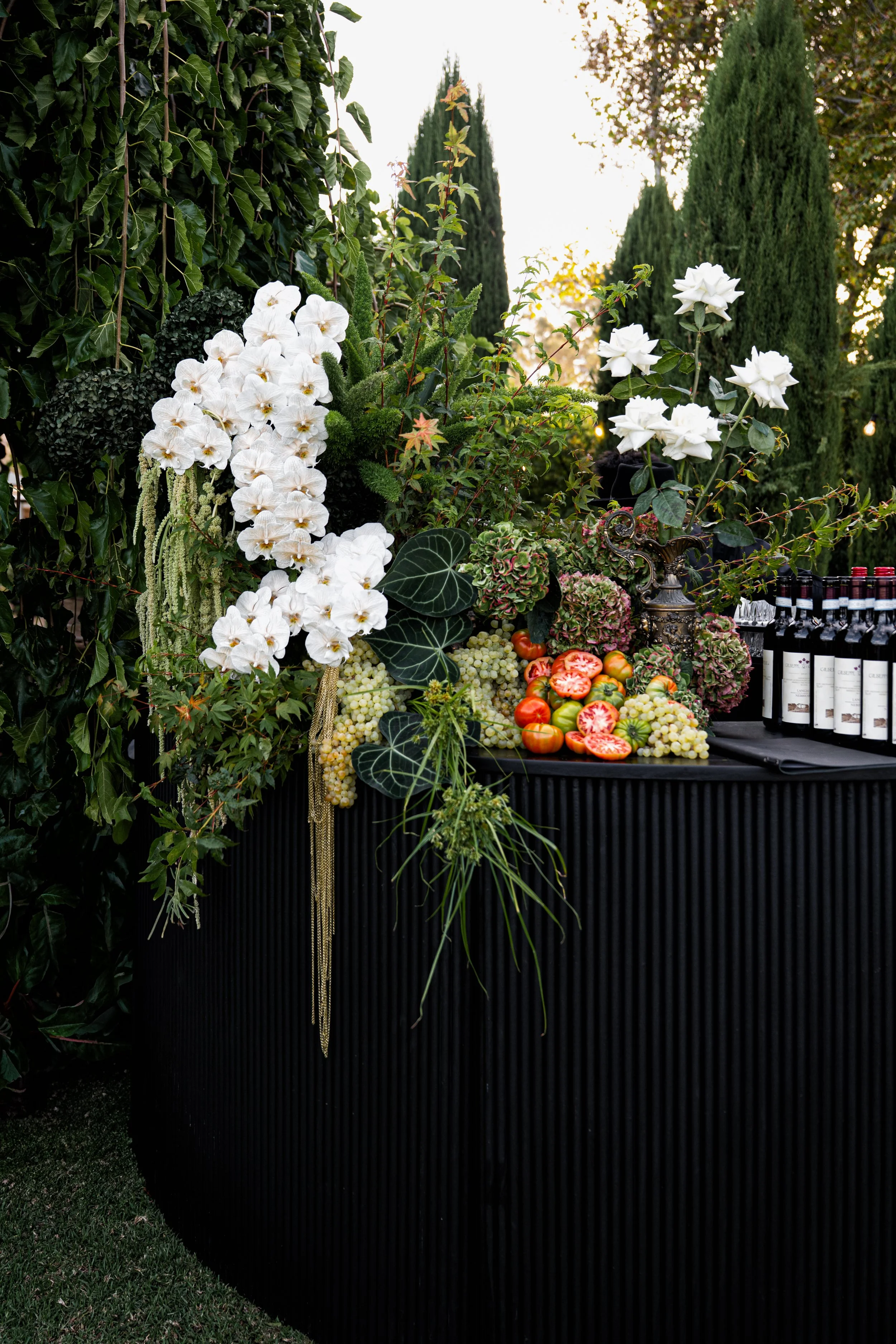 A black backdrop in front of a lush green hedge and tall cypress trees, decorated with an arrangement of white flowers including orchids and roses, as well as some tomatoes and grapes, and a display of wine bottles on the right.