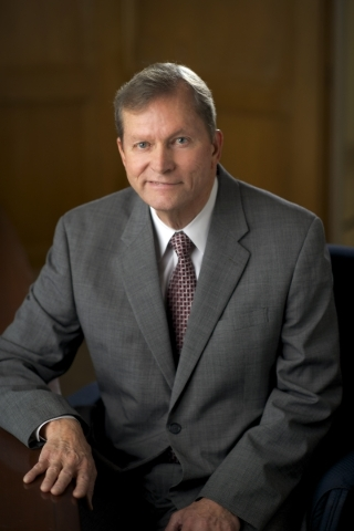 Professional man in a gray suit and tie, seated at a desk in a wood-paneled office, looking at the camera.