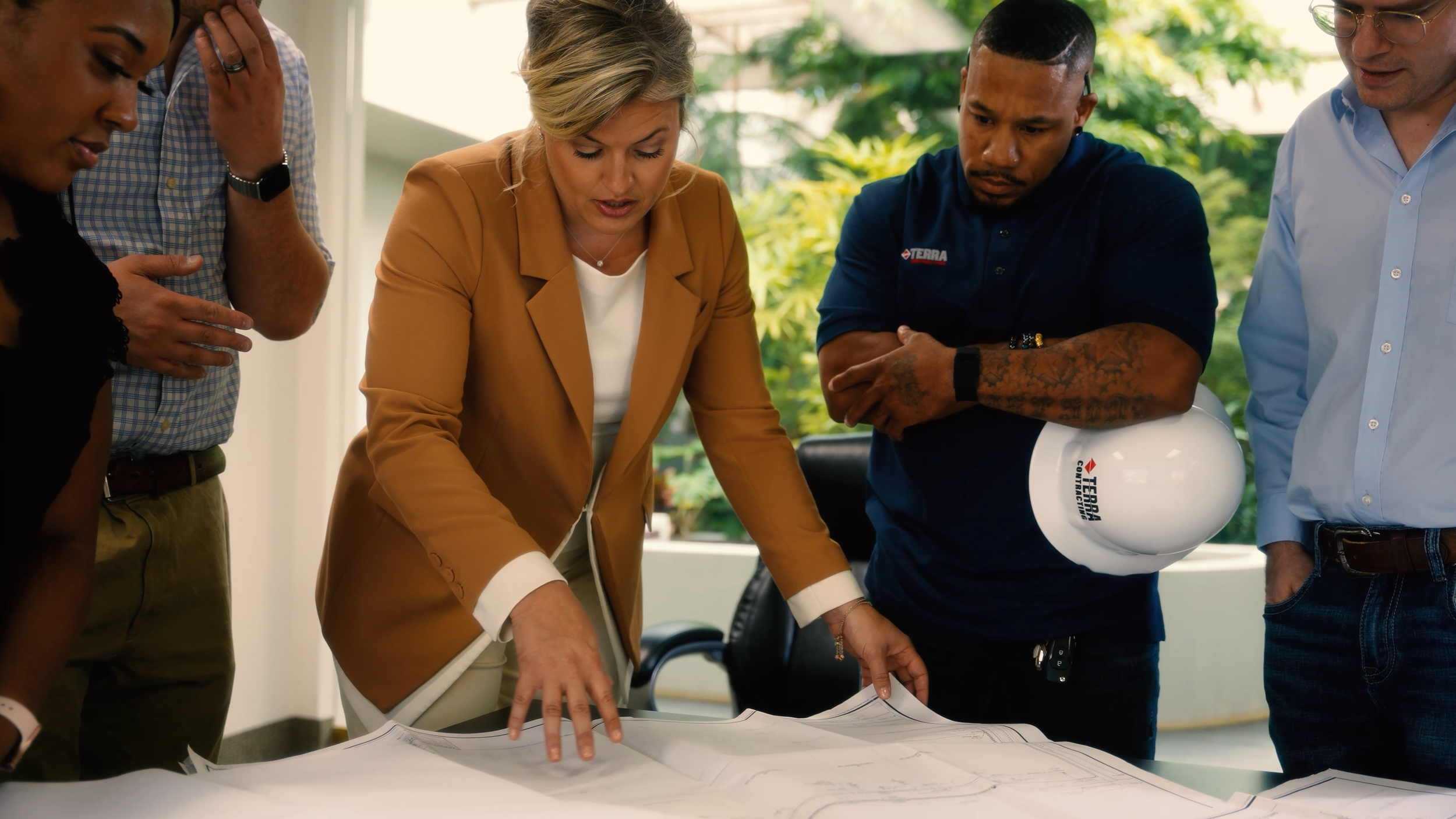 Group of professionals, including a woman in a tan blazer and a man holding a white safety helmet, gathered around a table looking at large blueprints during a discussion.