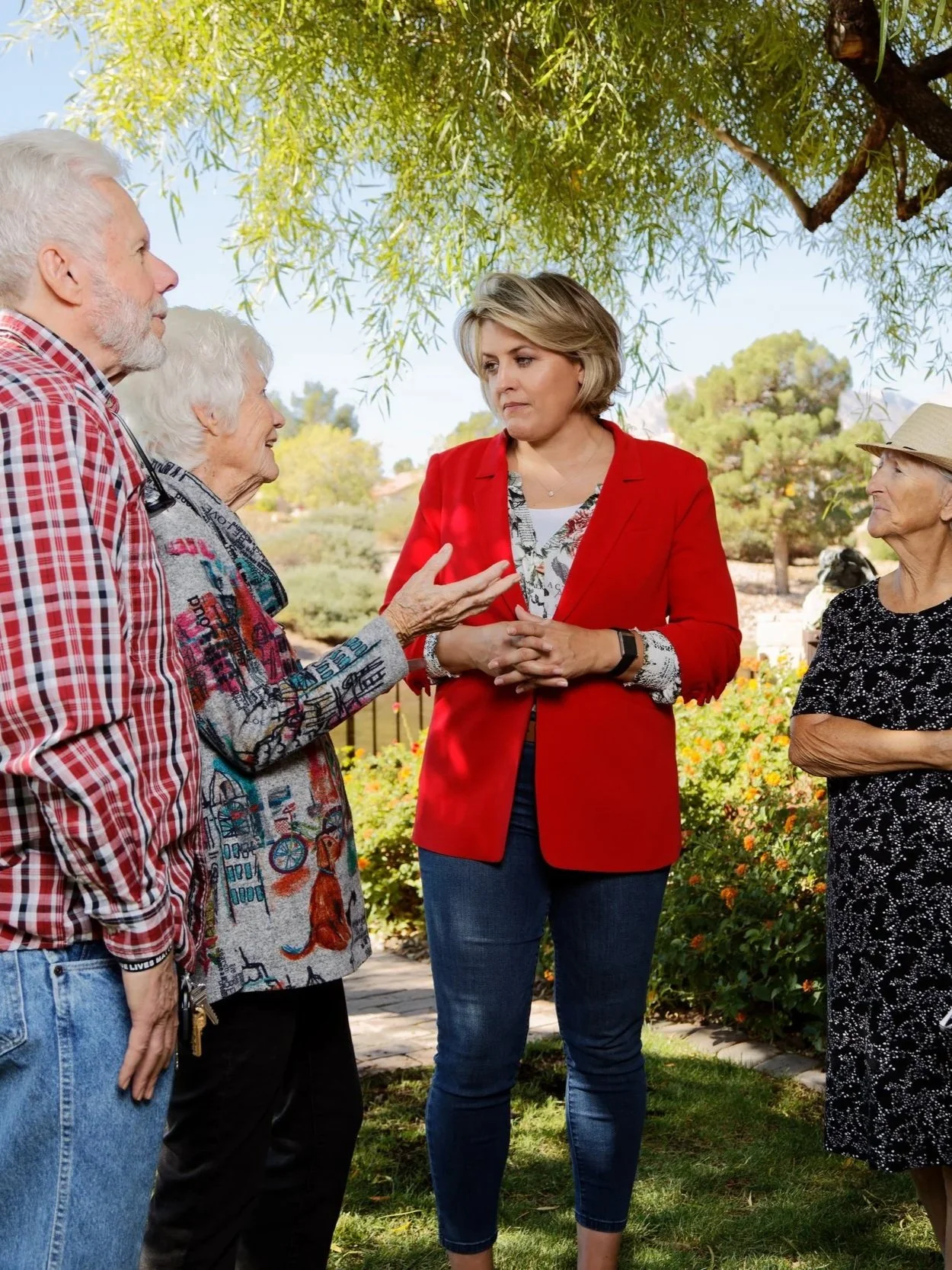 A woman in a red blazer speaking to a group of elderly people outdoors under a tree.
