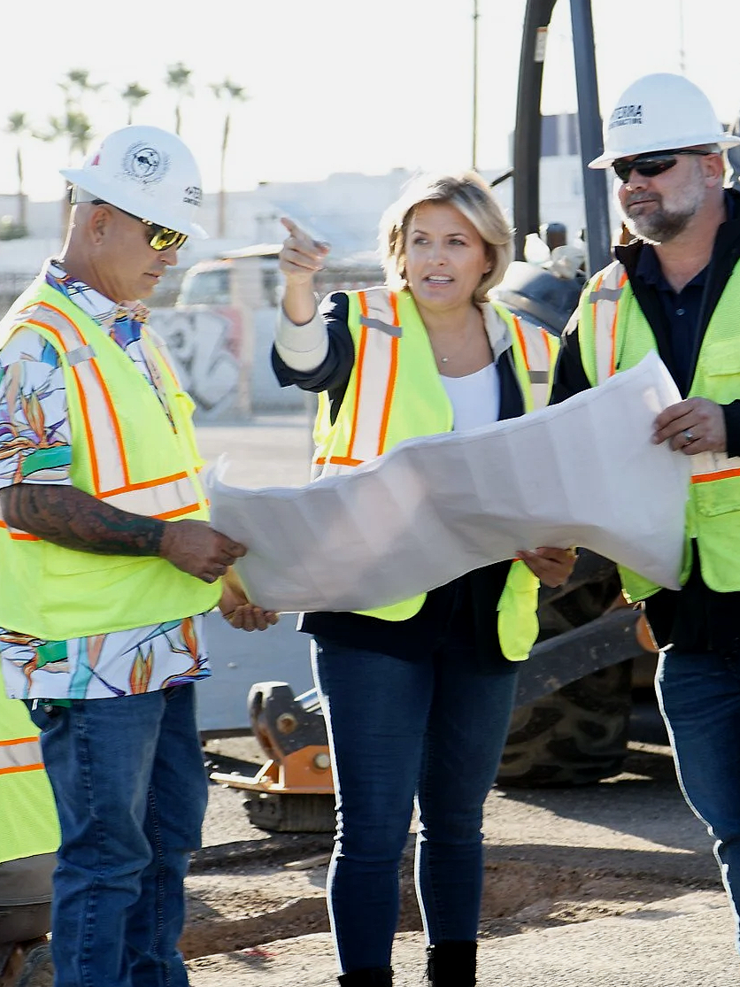 Three construction workers in safety vests and hard hats reviewing blueprints on a construction site with heavy machinery in the background.