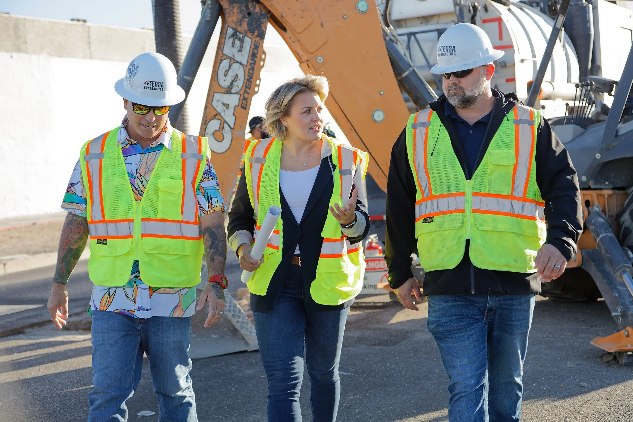 Three construction workers, two men and one woman, walking and talking on a construction site. They are wearing high-visibility vests, helmets, and sunglasses, with a large piece of construction machinery in the background.