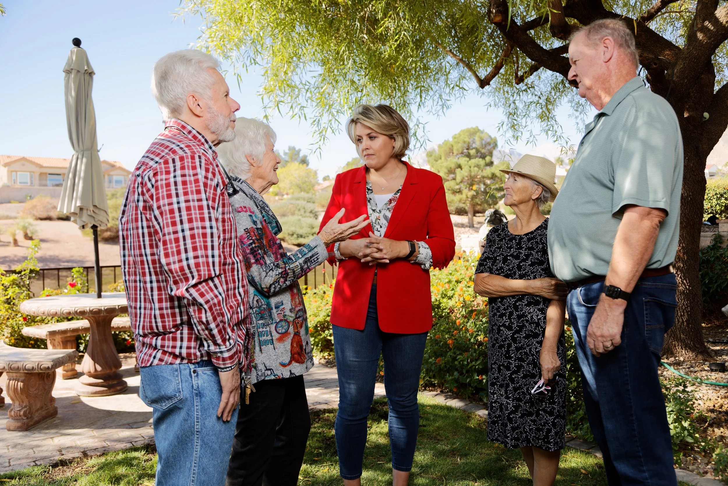 Six older adults and a woman in a red blazer engaged in conversation outdoors in a garden with trees, flowers, and a table with an umbrella.
