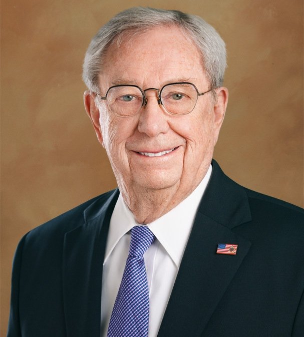 An older man with gray hair, glasses, wearing a dark suit, white shirt, and a light purple tie, smiling in a professional portrait.