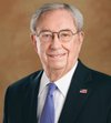 An older man with gray hair, glasses, wearing a dark suit, white shirt, and a light purple tie, smiling in a professional portrait.
