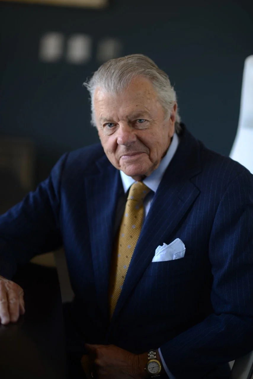 An older man with gray hair and blue eyes dressed in a navy pinstripe suit with a white shirt, yellow tie, and white pocket square, sitting at a desk in an office environment.