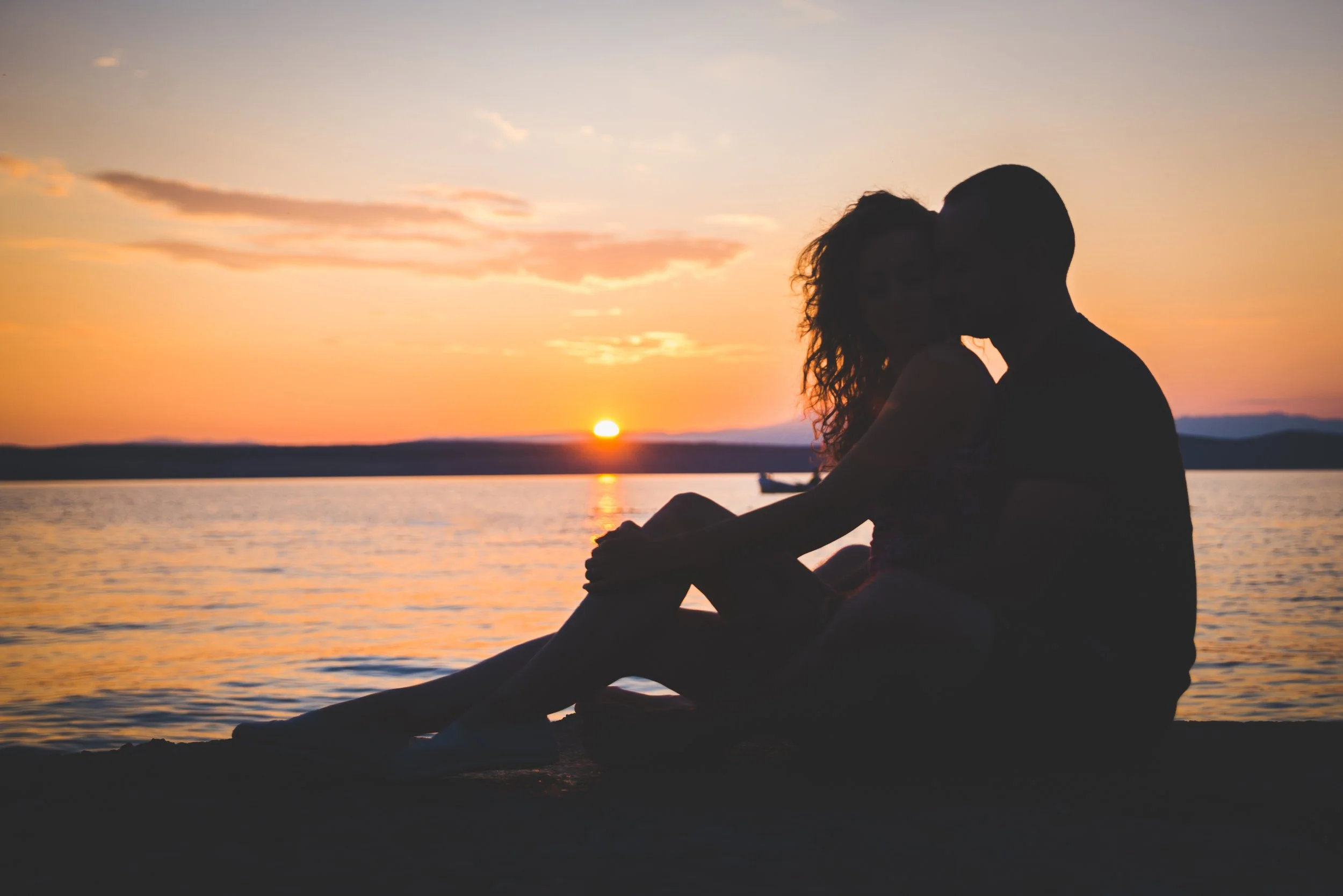 A couple sitting by the water during sunset, with the man kissing the woman's forehead.