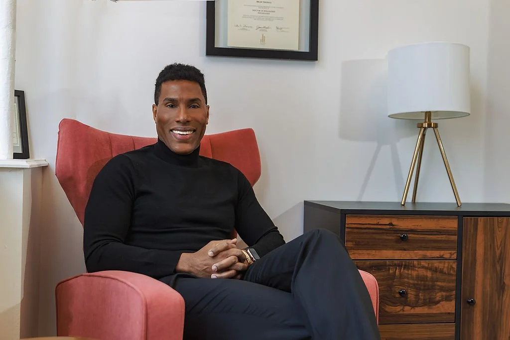 A man sitting in a pink armchair in an office, smiling at the camera. There is a framed certificate or diploma on the wall behind him, a lamp on a wooden cabinet to his right, and a window with a white curtain on his left.