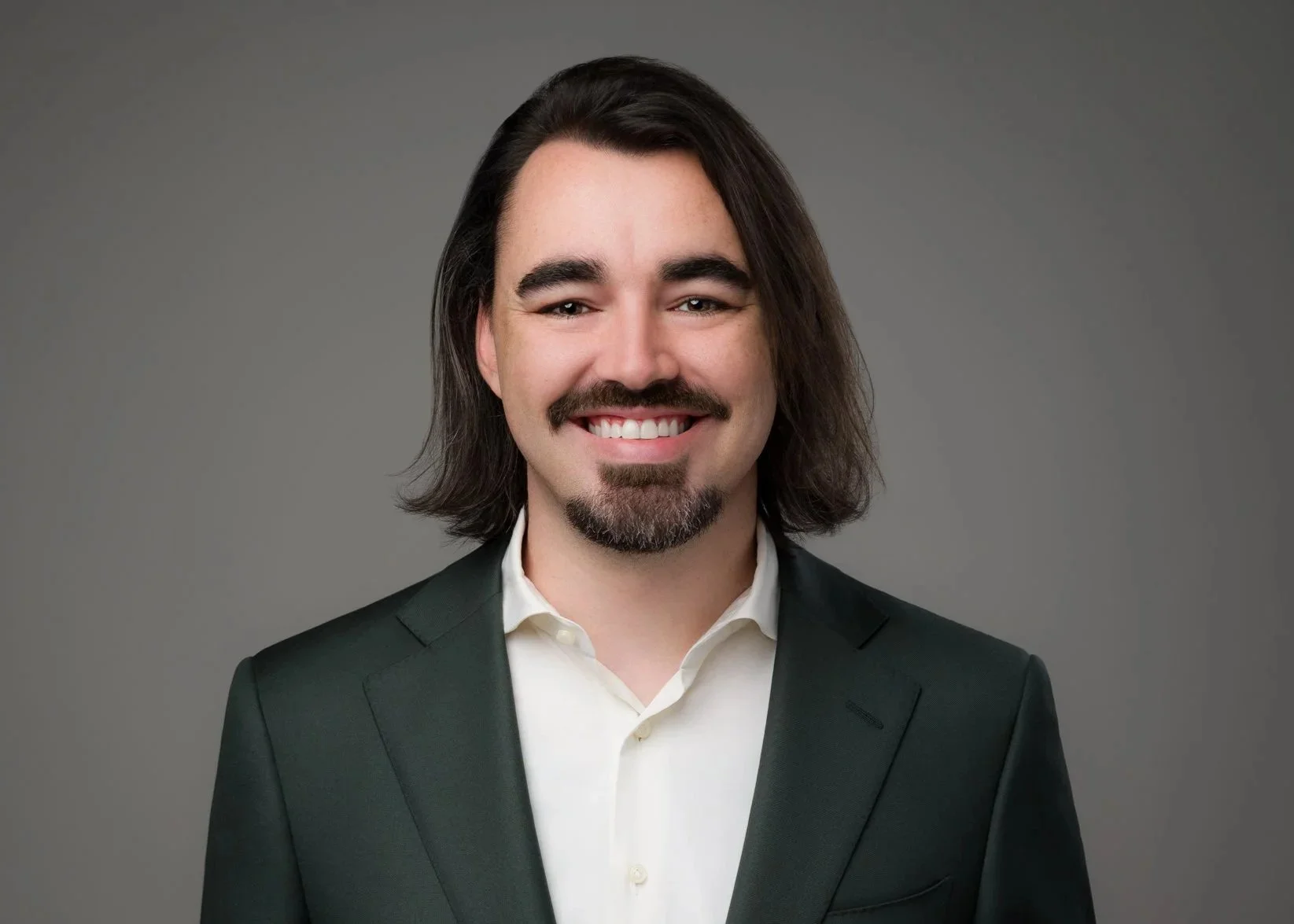A man with long dark hair, a mustache, and goatee, wearing a dark suit jacket and white shirt, smiling against a gray background.