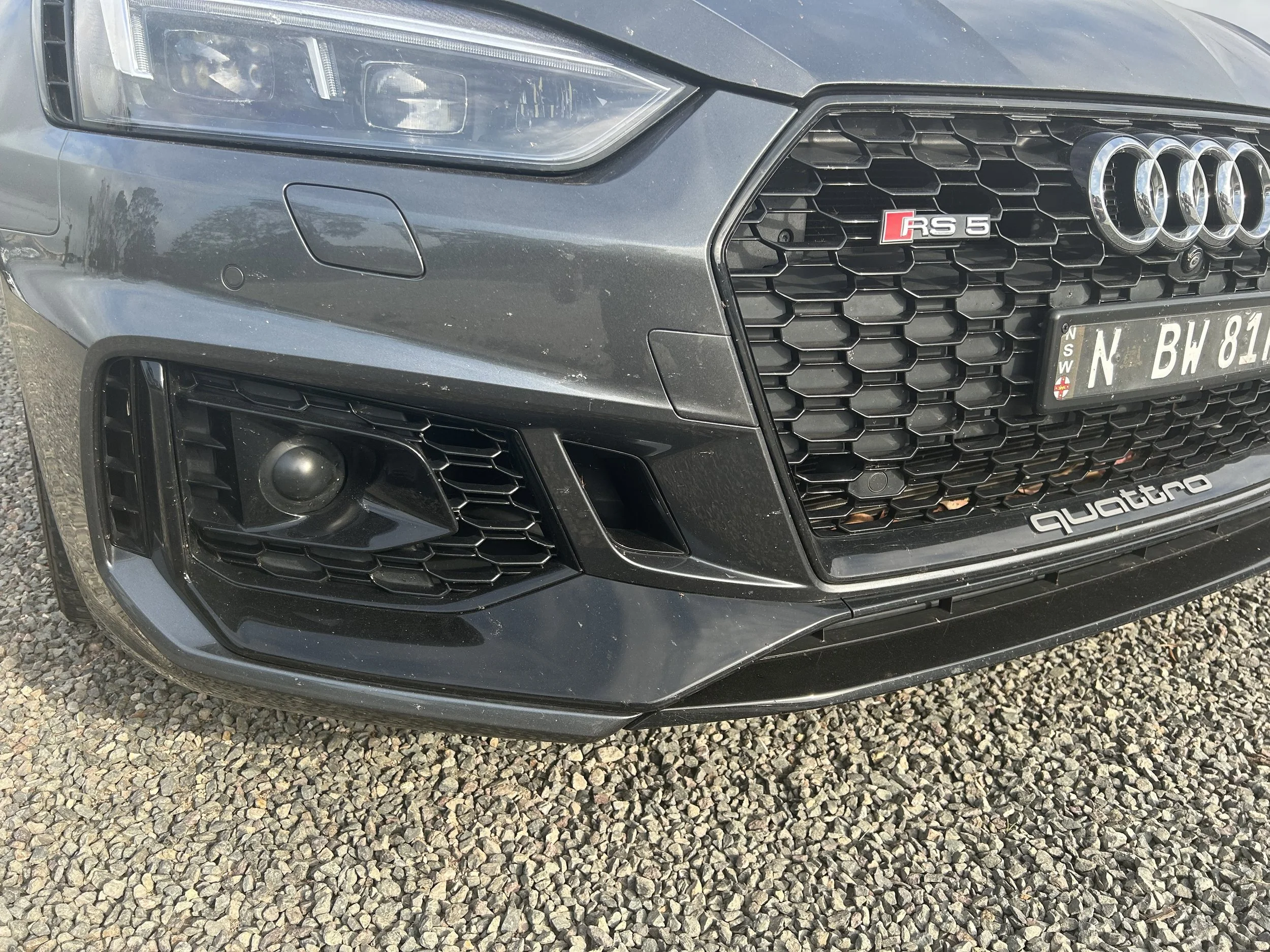 Close-up of the front bumper of a grey Audi RS5 before detailing, showing dirt, scuff marks, and imperfections on the grille, headlights, and lower bumper.