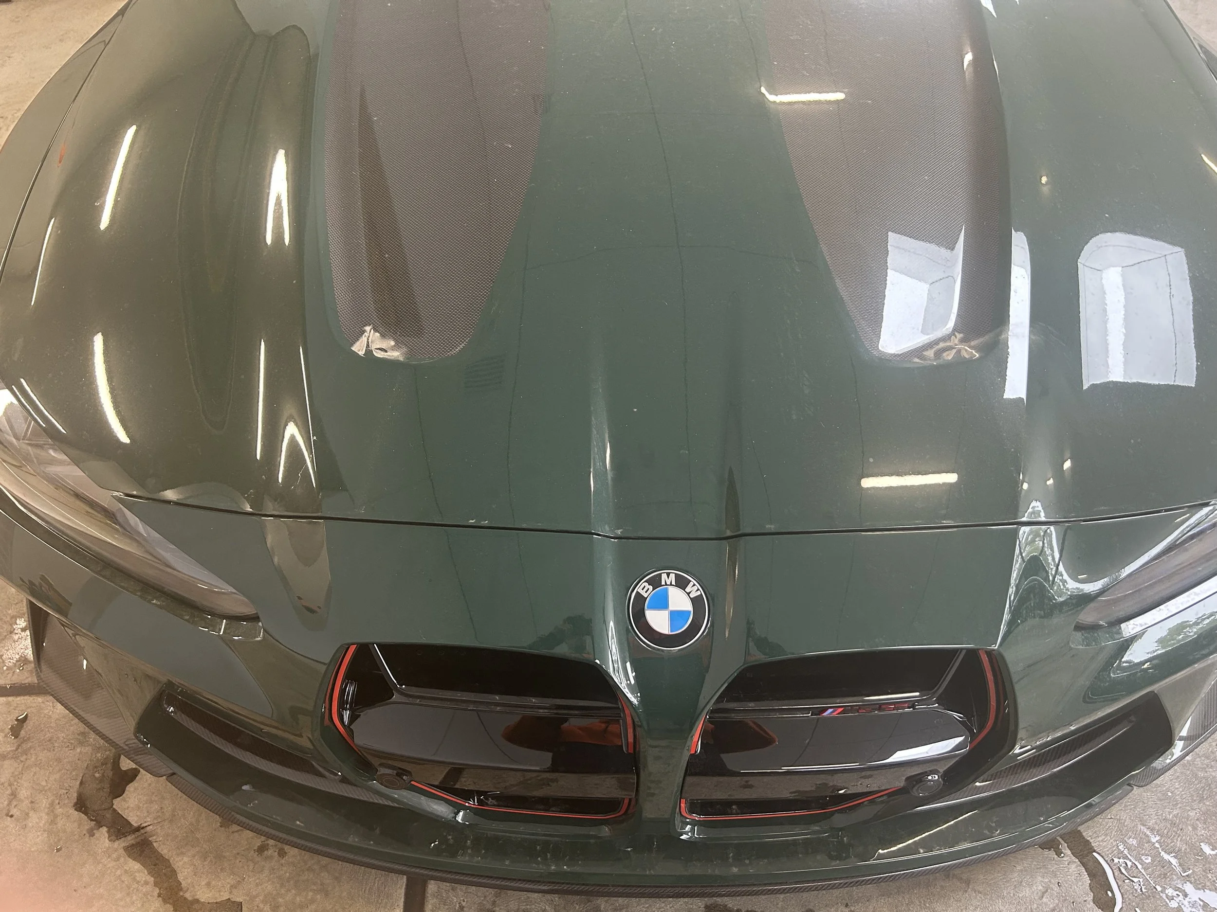 Top view of a dark green BMW sports car with a split hood, showing the BMW logo at the front center and the black grille with red accent trim. The car has two large windshield vents and reflections of overhead lights on the hood.