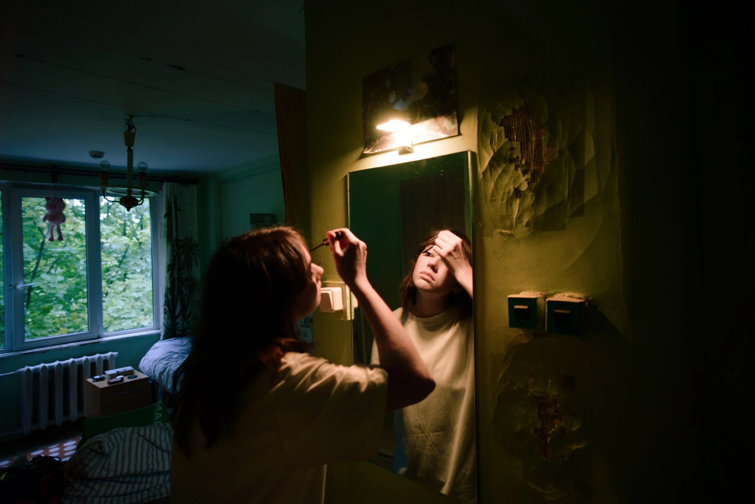 Young woman looking into a mirror in a dark apartment bathroom in Russia. Photograph by Semyon Kokorin featured in “Semyon’s World” by Timofey Abel.