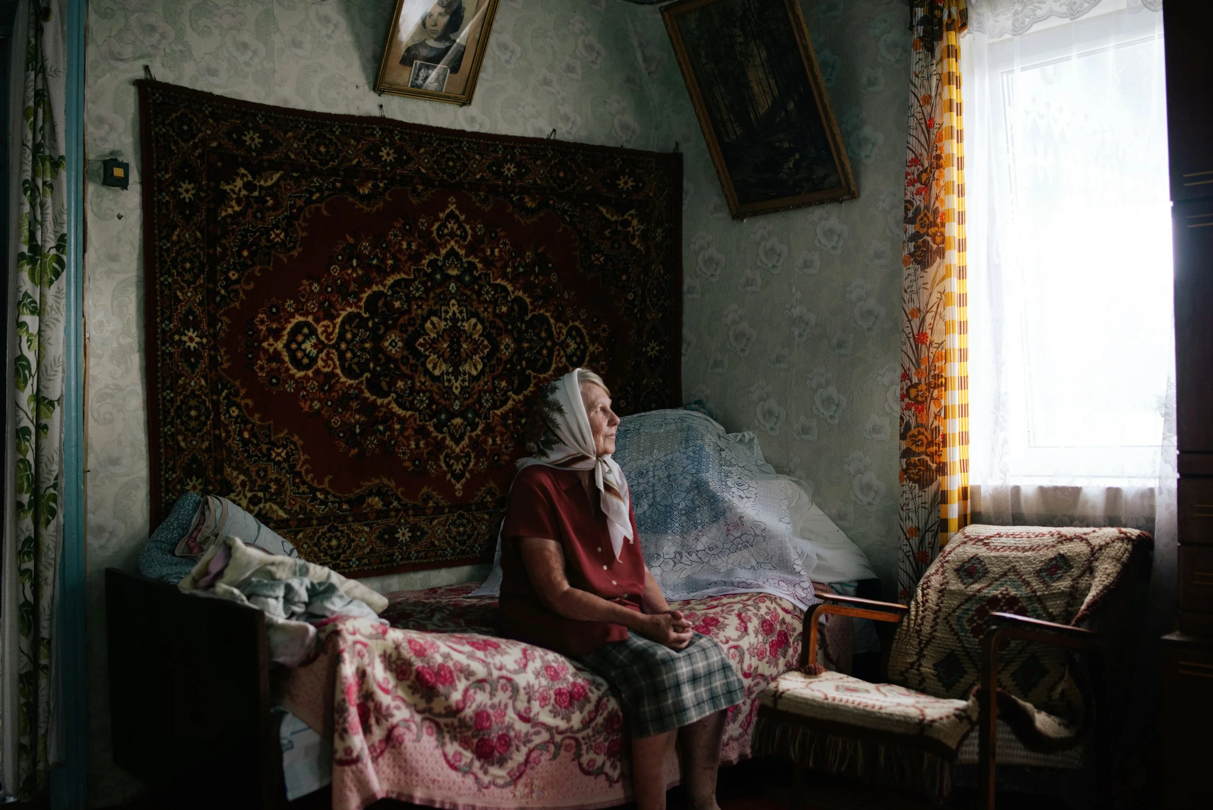 Elderly woman sitting in a modest bedroom with Soviet-era interior details in rural Russia. Photograph by Semyon Kokorin featured in “Semyon’s World” by Timofey Abel.