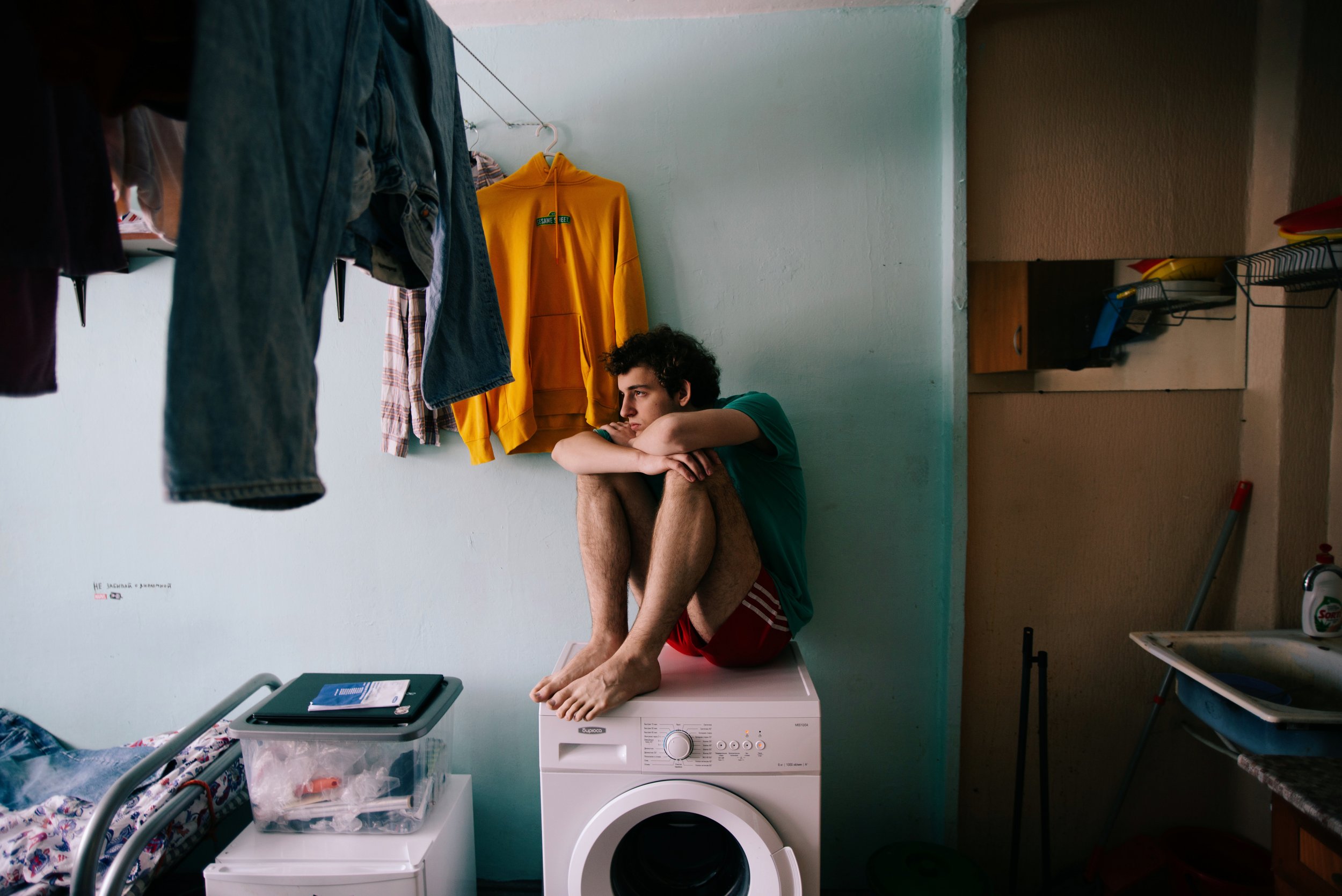 Young man sitting on a washing machine inside a small apartment in Russia. Photograph by Semyon Kokorin featured in “Semyon’s World” by Timofey Abel.