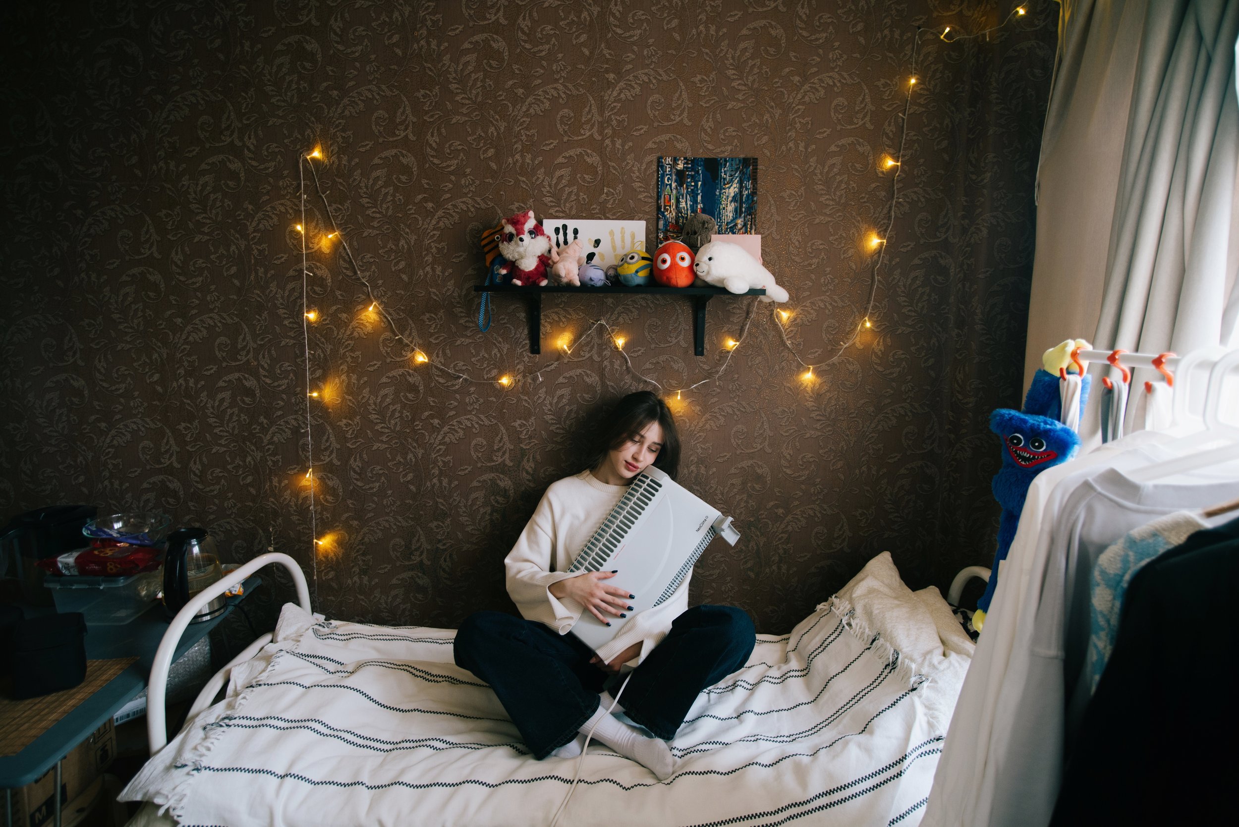 Young woman sitting on a bed holding a keyboard in a dimly lit bedroom in Russia. Photograph by Semyon Kokorin featured in “Semyon’s World” by Timofey Abel.