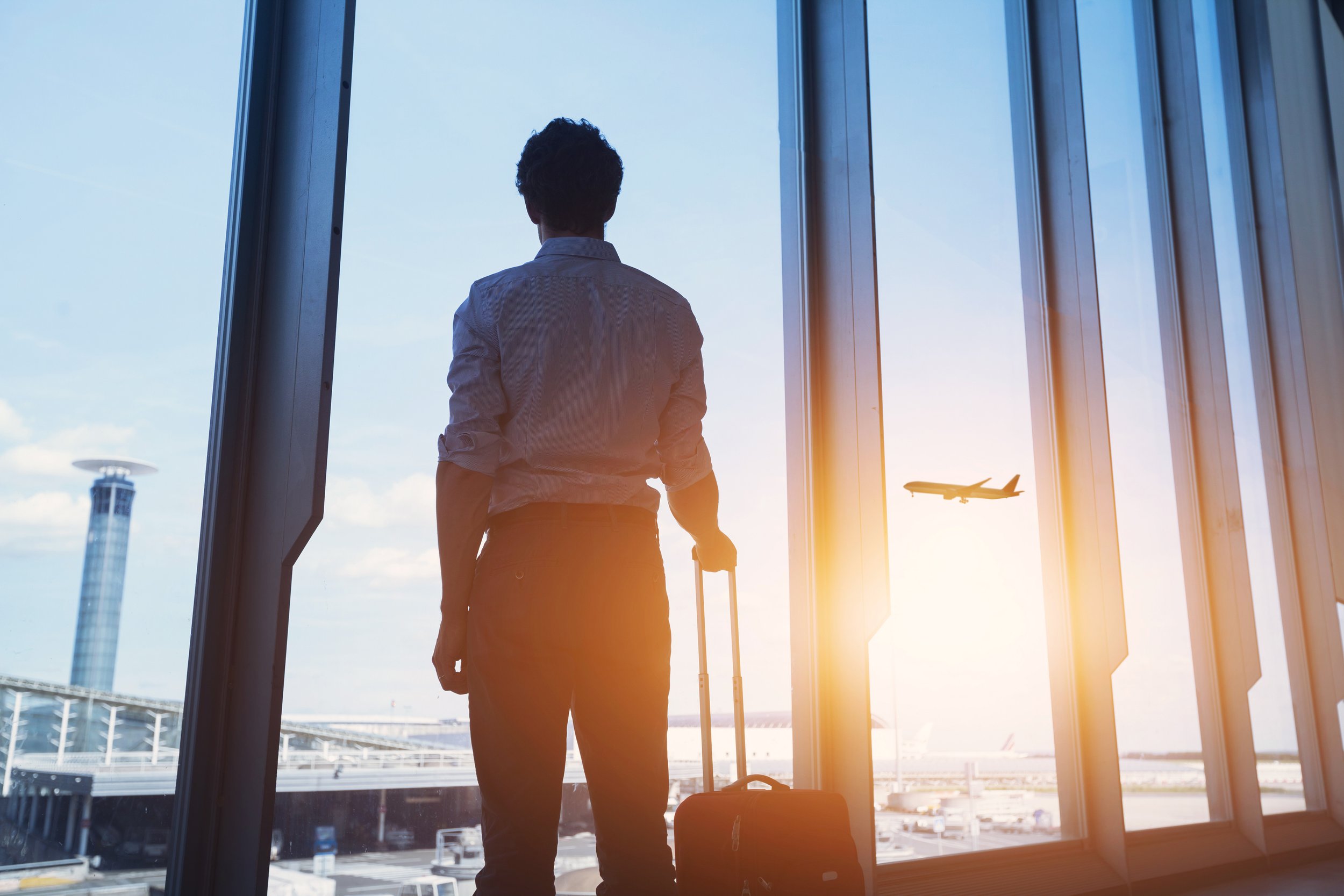 A person with a rolling suitcase standing at an airport terminal, looking out at an airplane flying in the sky during sunset.