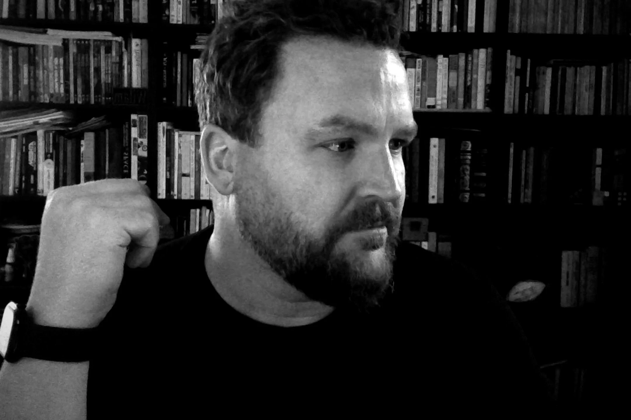 A man with a beard and short hair, wearing a black shirt, adjusting his sleeve, with a background of bookshelves filled with books.