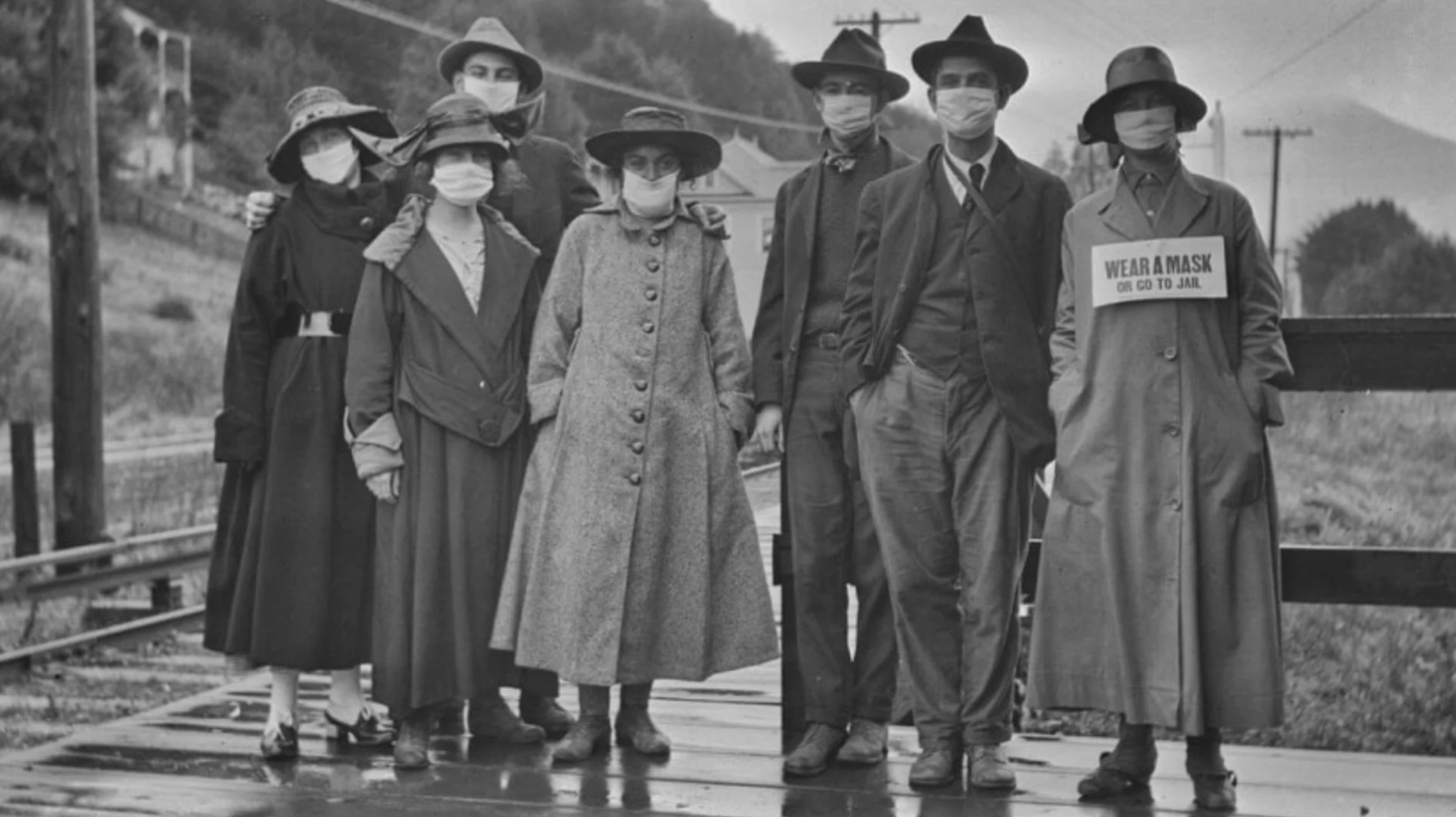 Black and white photo of a group of people wearing masks, standing outdoors on a street in Utah during the Spanish Flu epidemic of 1918. They are dressed in vintage clothing, and one person is holding a sign that reads, 'Wear a Mask or Go to Jail.'