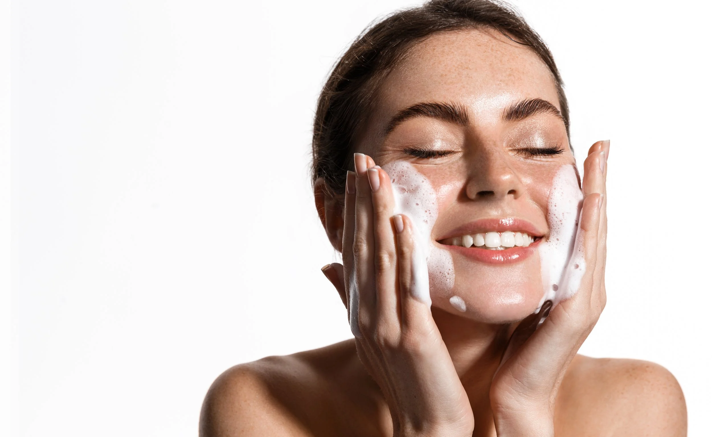 A woman with brown hair washing her face with foamy soap, smiling with eyes closed.
