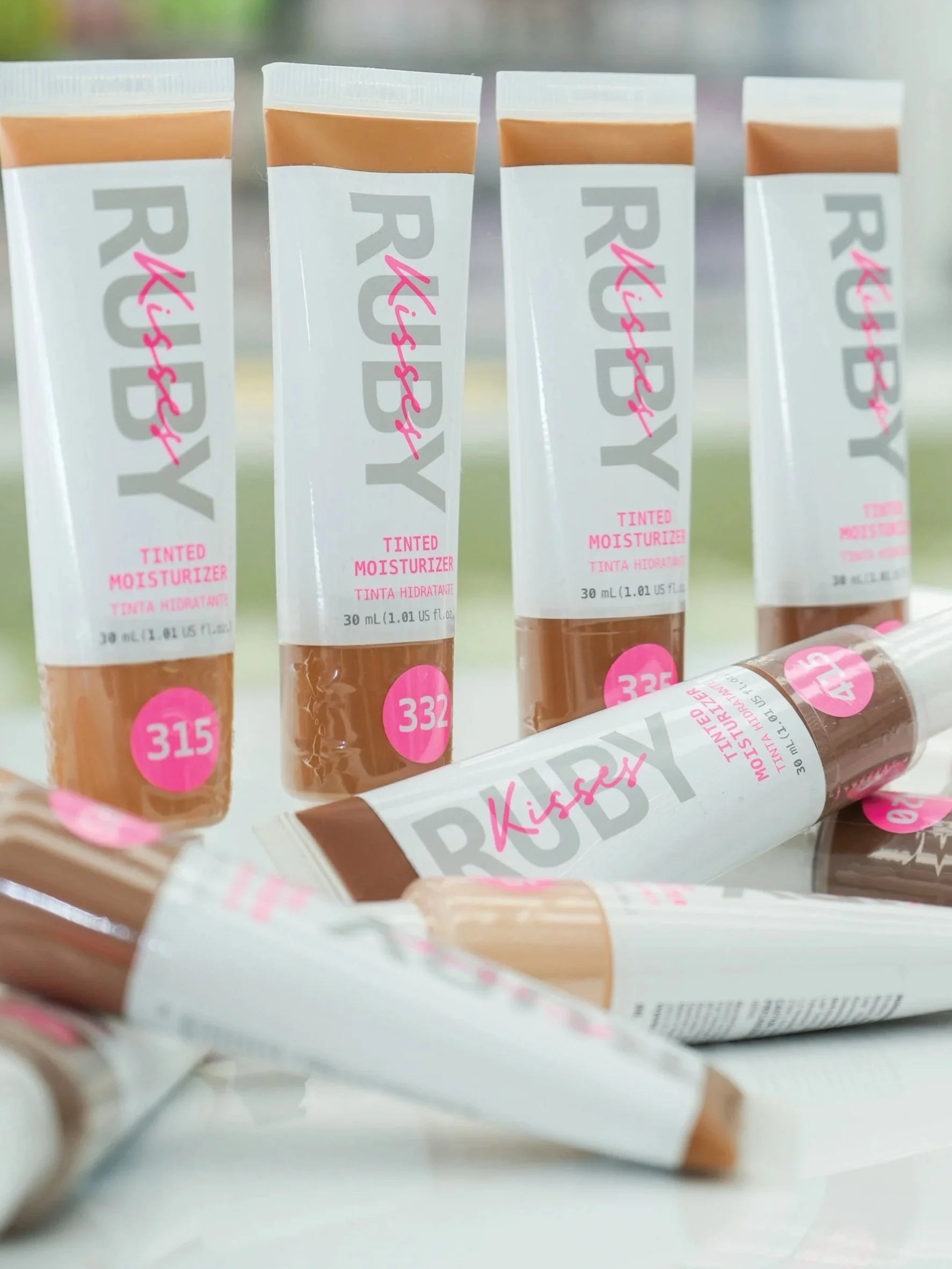 Multiple tubes of RIY Kidssky tinted moisturizer, with some lying on a white surface and others standing upright. Each tube is 30 mL and features a beige cap, pink circles with product numbers, and pink text on the packaging.