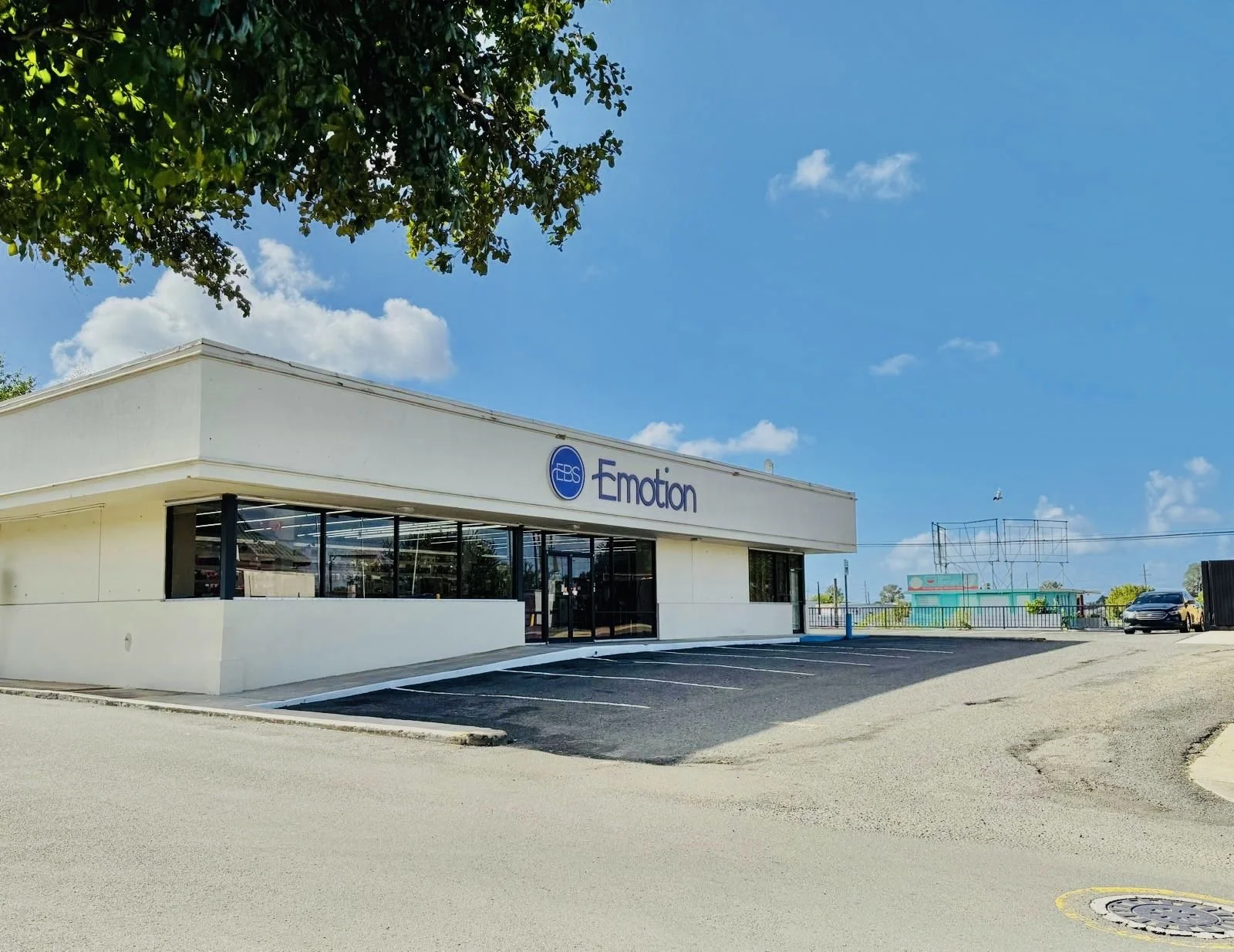 A white commercial building with large glass windows and a blue sign that reads 'EBS' and 'Emotion'. There is a parking lot with spaces and a few cars parked. The sky is bright blue with scattered clouds and trees are visible in the background.