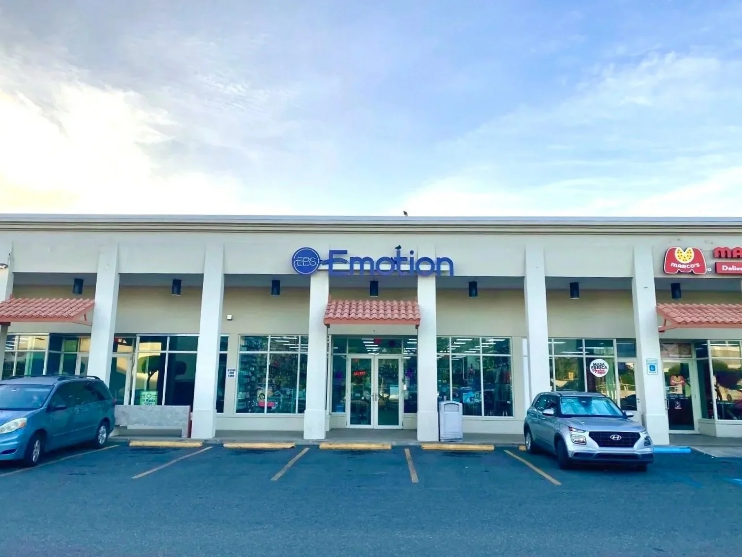Front view of a shopping plaza with parking lot, featuring a store called 'Elation' with a blue sign, and other stores partially visible on the right, under a partly cloudy sky.