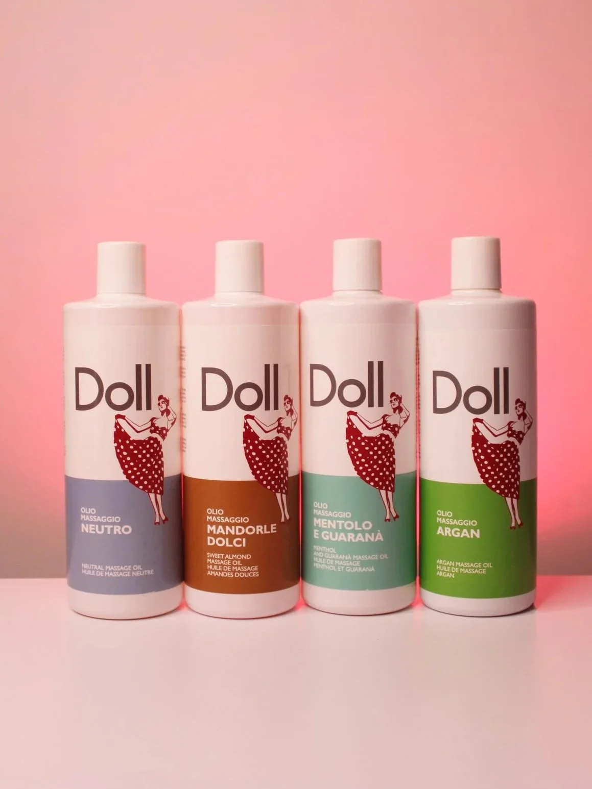 Four bottles of Doll massage oil in different scents: neutral, sweet almond, mint and guarana, and argan, lined up on a pink surface against a pink background.