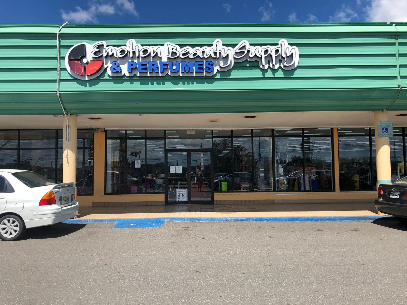 Storefront with a sign reading 'Emotion Beauty Supply & Perfumes' in front of a parking lot with a disabled parking space and cars.