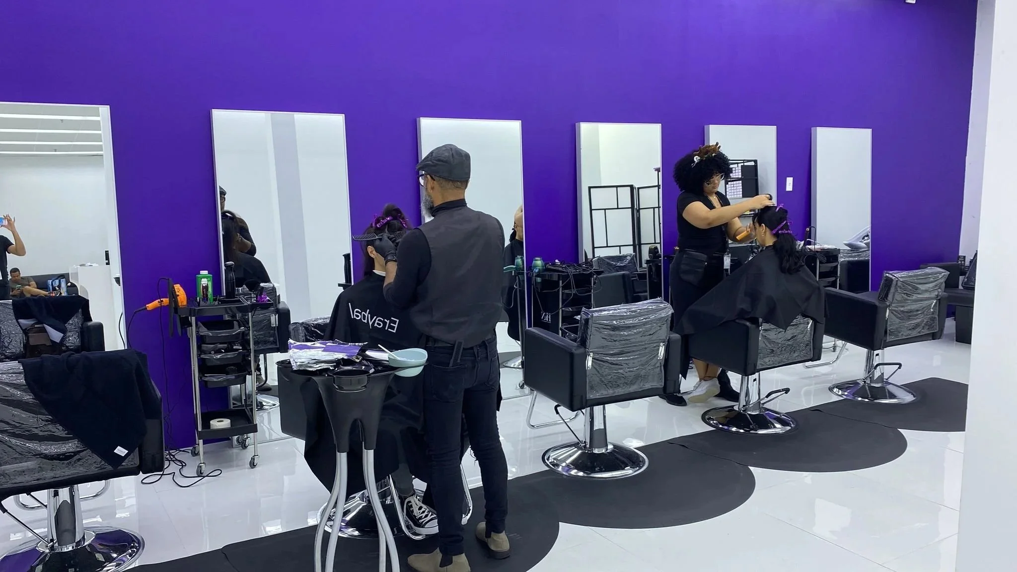 A hair salon with purple walls and black styling chairs. Two hairstylists are working on clients' hair, with mirrors and styling tools visible.