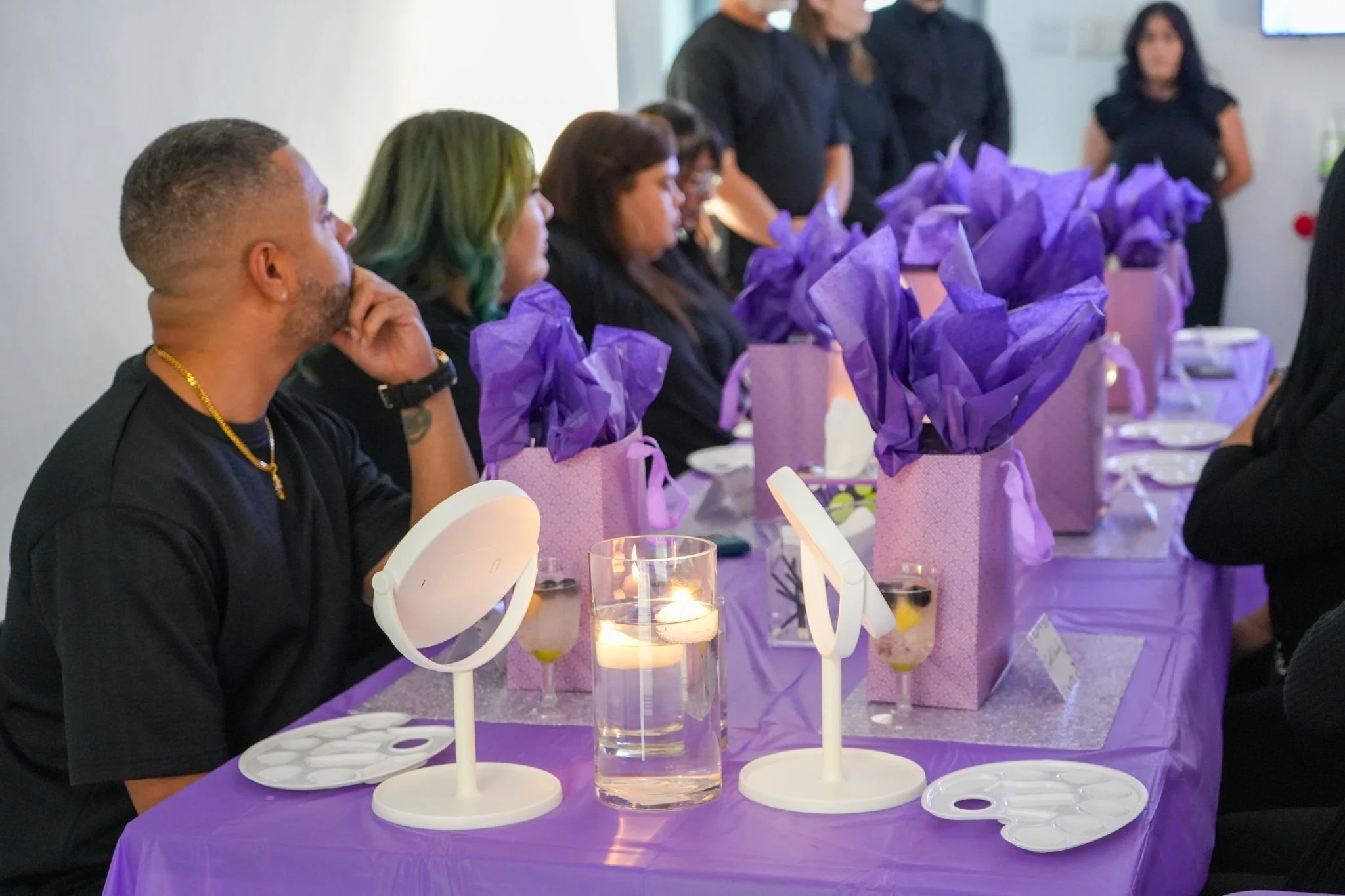 People gathered around a purple-themed table for a celebration or event, with purple gift bags, tissue paper, and purple decorations, while someone appears to be getting their makeup done.