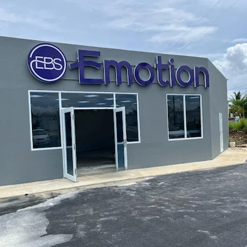 Exterior of a gray commercial building with a sign that says 'EBS Emotion' and an open glass door. The building has large windows and is situated on a paved lot with a cloudy sky visible in the background.
