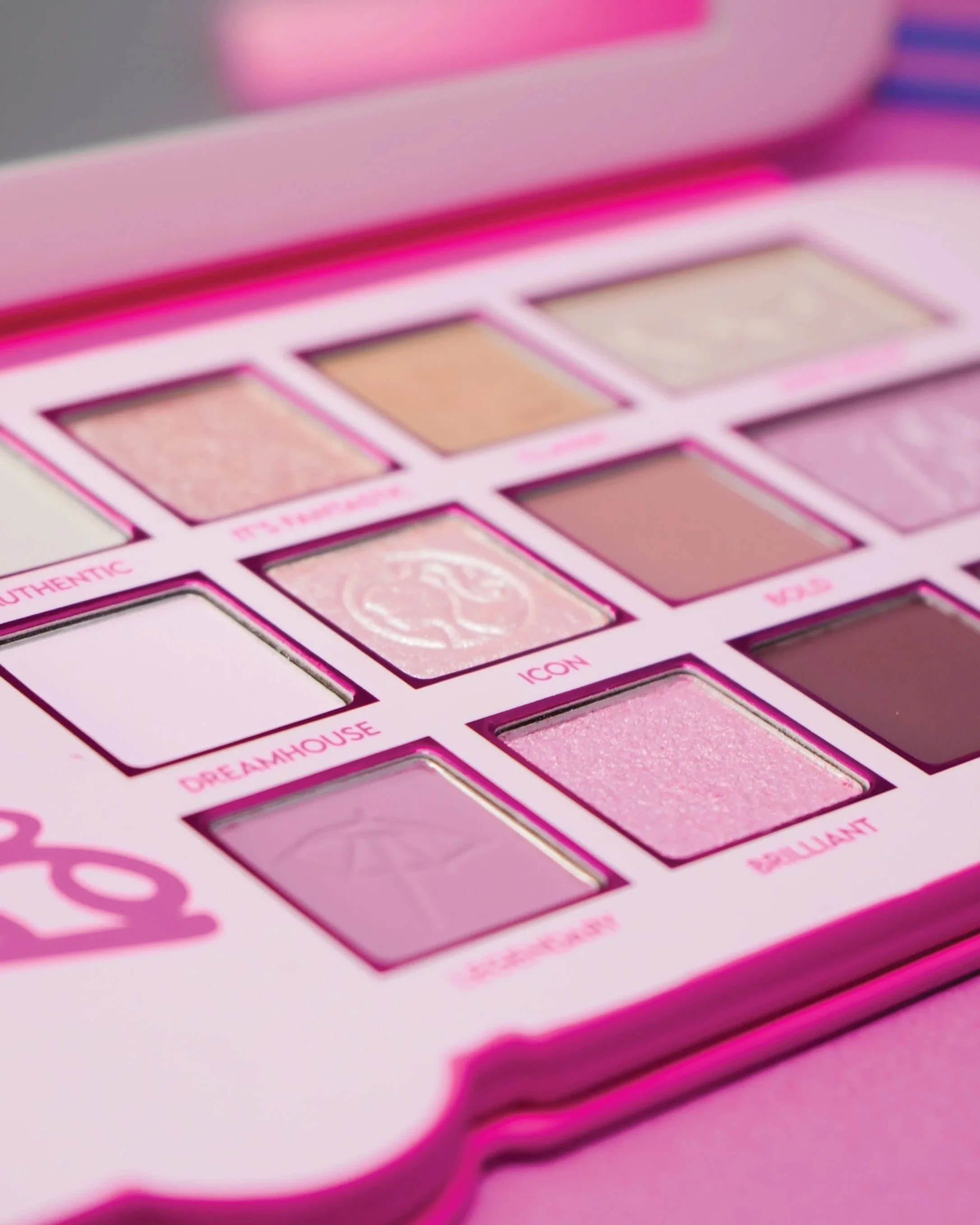 A pink eyeshadow palette with multiple shades, including shimmery and matte colors, arranged in rows with labeled sections such as 'Authentic,' 'Dreamhouse,' 'Icon,' and 'Brilliant.'