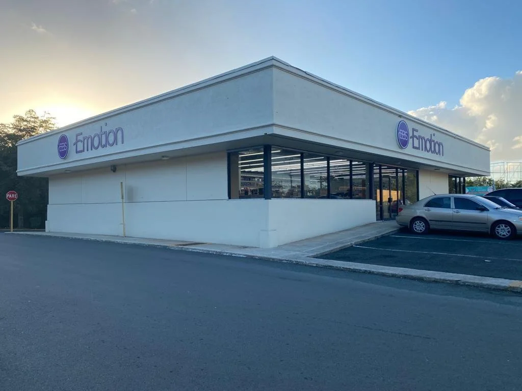 A white commercial building with glass windows displaying the store name 'Emoción' and a purple logo, with a parking lot and cars in front, under a partly cloudy sky.