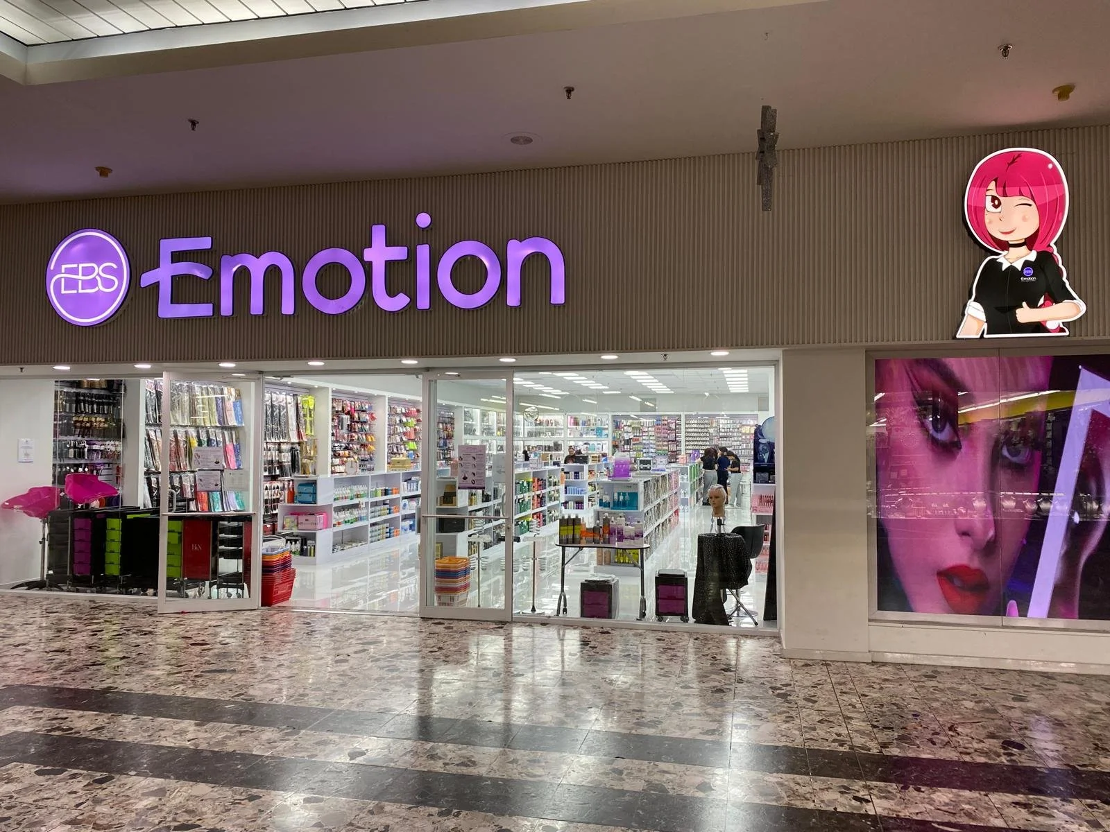 Entrance of a store named 'Emotion' with purple signage, inside shelves filled with stationery and cosmetic products, and large promotional images of a woman with pink hair and makeup.