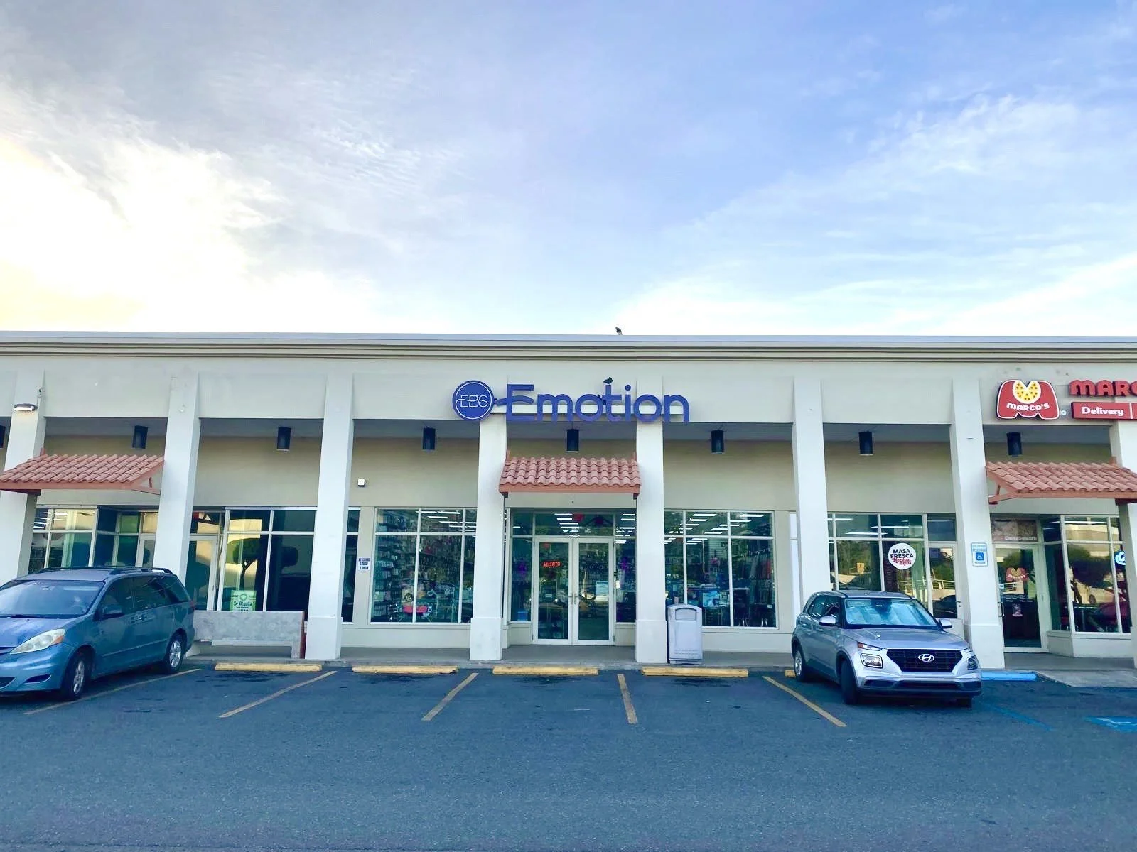 Shopping plaza with stores including ECS Emotion, Marco's pizza delivery, and other shops, with parking lot in front and a blue sky above.