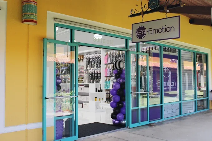 Entrance to a store named EBS Emotion with glass doors and windows, decorated with purple and black balloons, inside are shelves with products, located in a shopping strip with a yellow exterior wall.